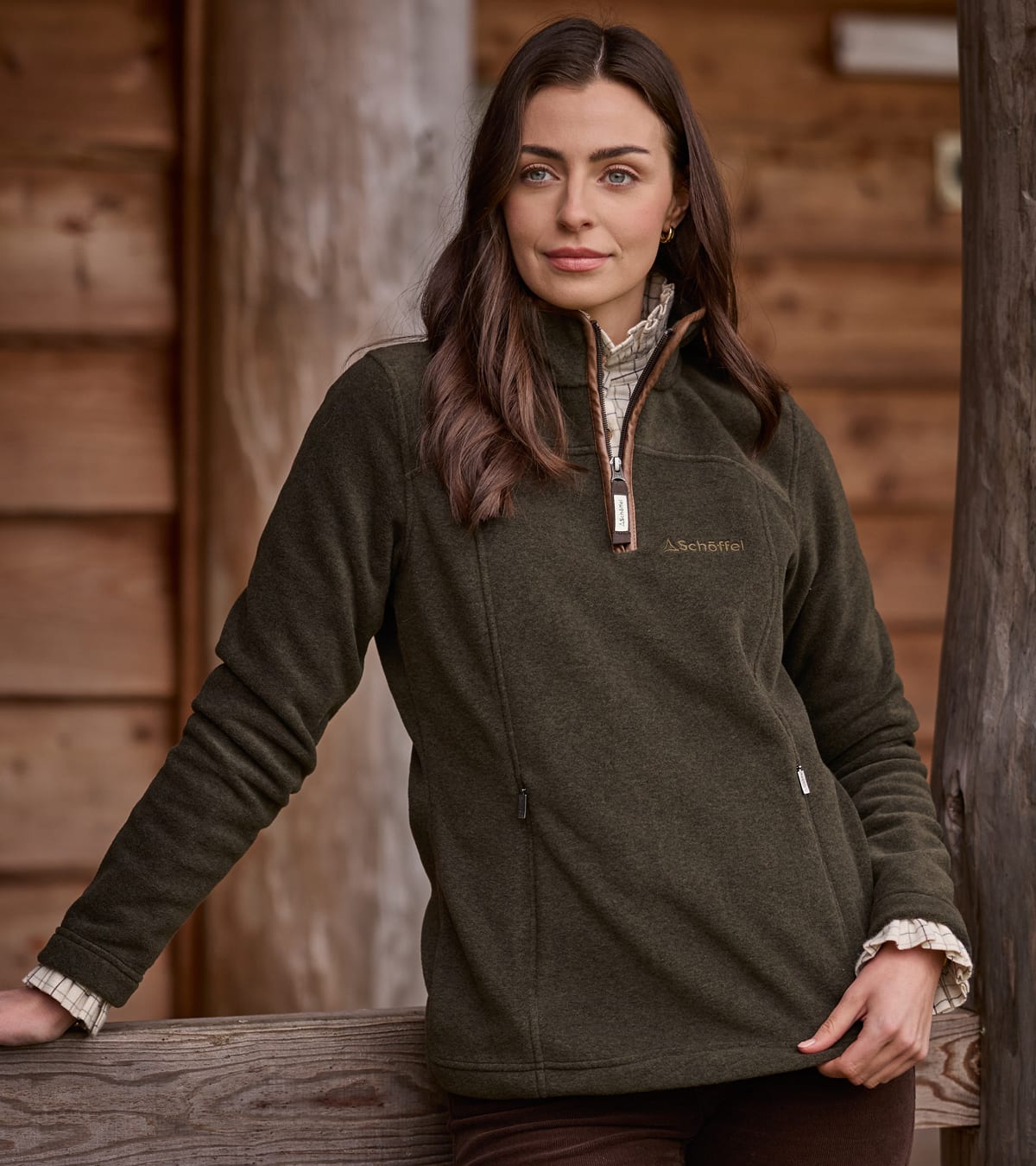 A woman wearing an olive green fleece with a quarter-zip posed outdoors against a rustic wooden wall.