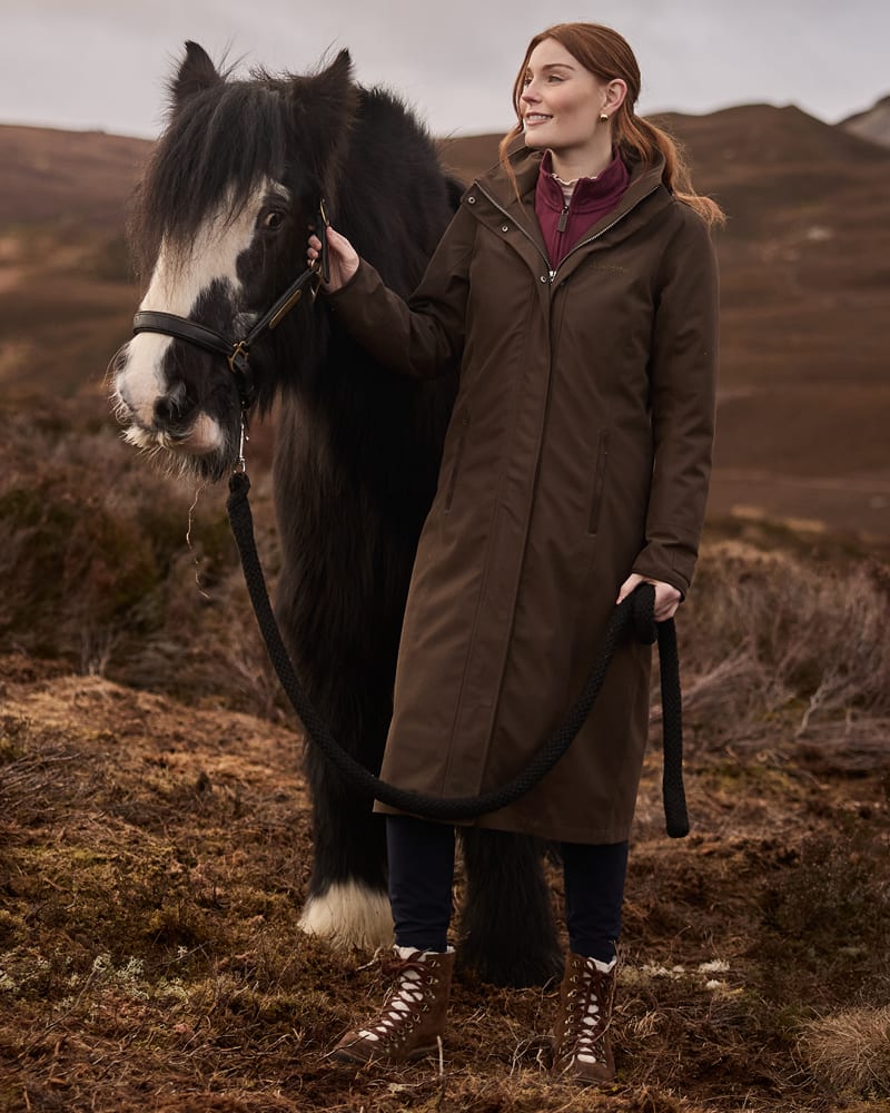 A woman stands outdoors with a horse, wearing a long brown coat over a burgundy top and dark pants. The background shows a natural, hilly landscape under an overcast sky.