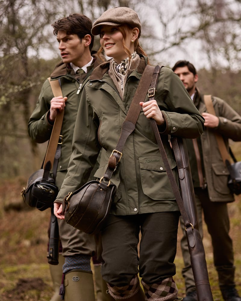 Three people outdoors in a wooded setting wearing matching olive green shooting jackets and carrying brown leather bags and rifles.