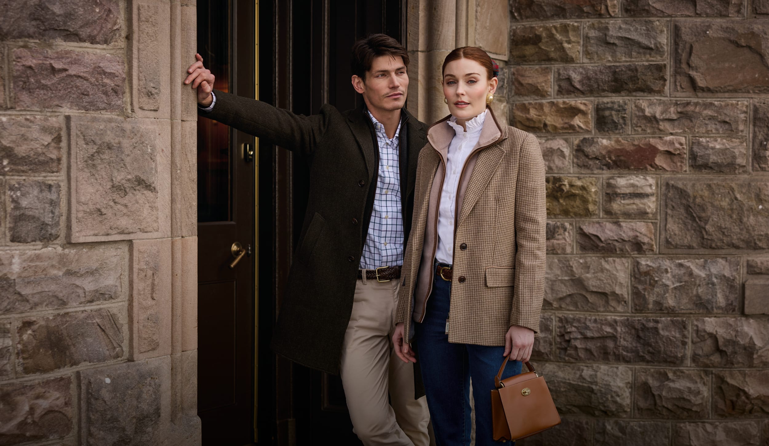 A man and a woman stand outside a stone building doorway. The man wears a dark overcoat over a light check shirt and beige trousers, while the woman wears a beige coat over a white top and blue jeans and carries a brown handbag.