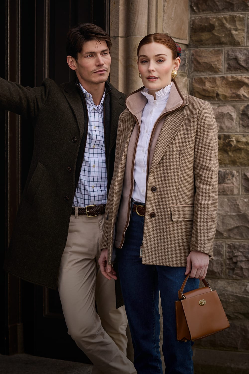A man and a woman pose outdoors in front of a stone wall. The woman wears a beige houndstooth blazer over a white ruffled blouse, blue jeans, and holds a brown leather handbag.