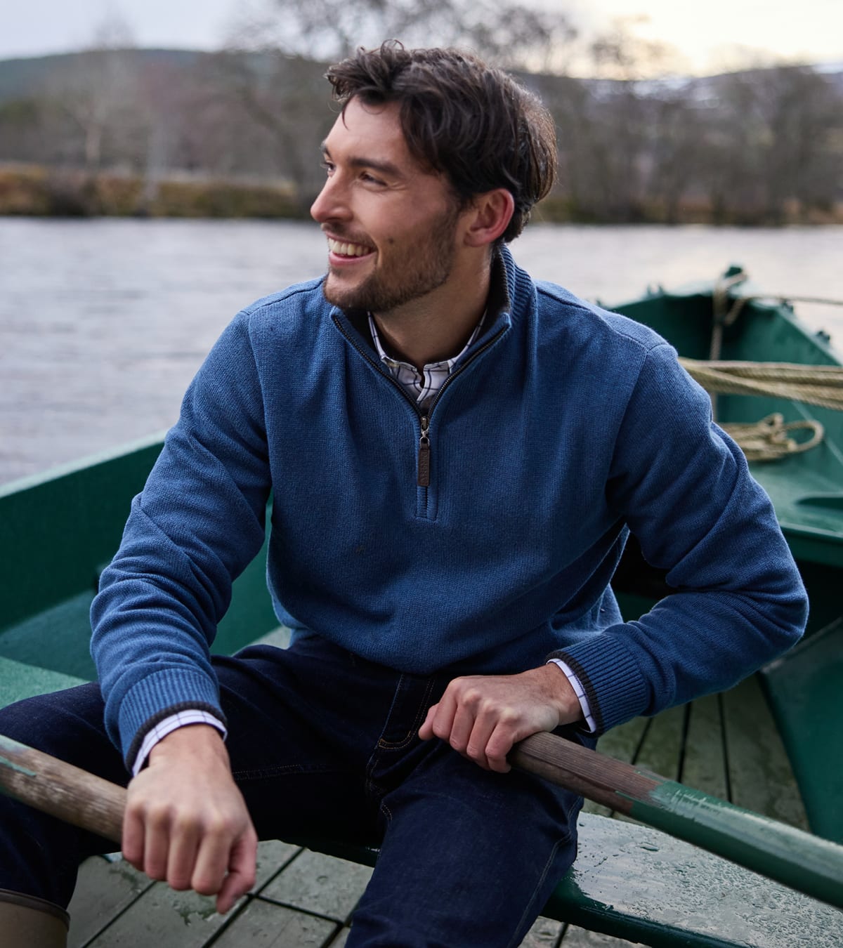 Man wearing blue quarter-zip knit jumper rowing a green wooden boat on a loch