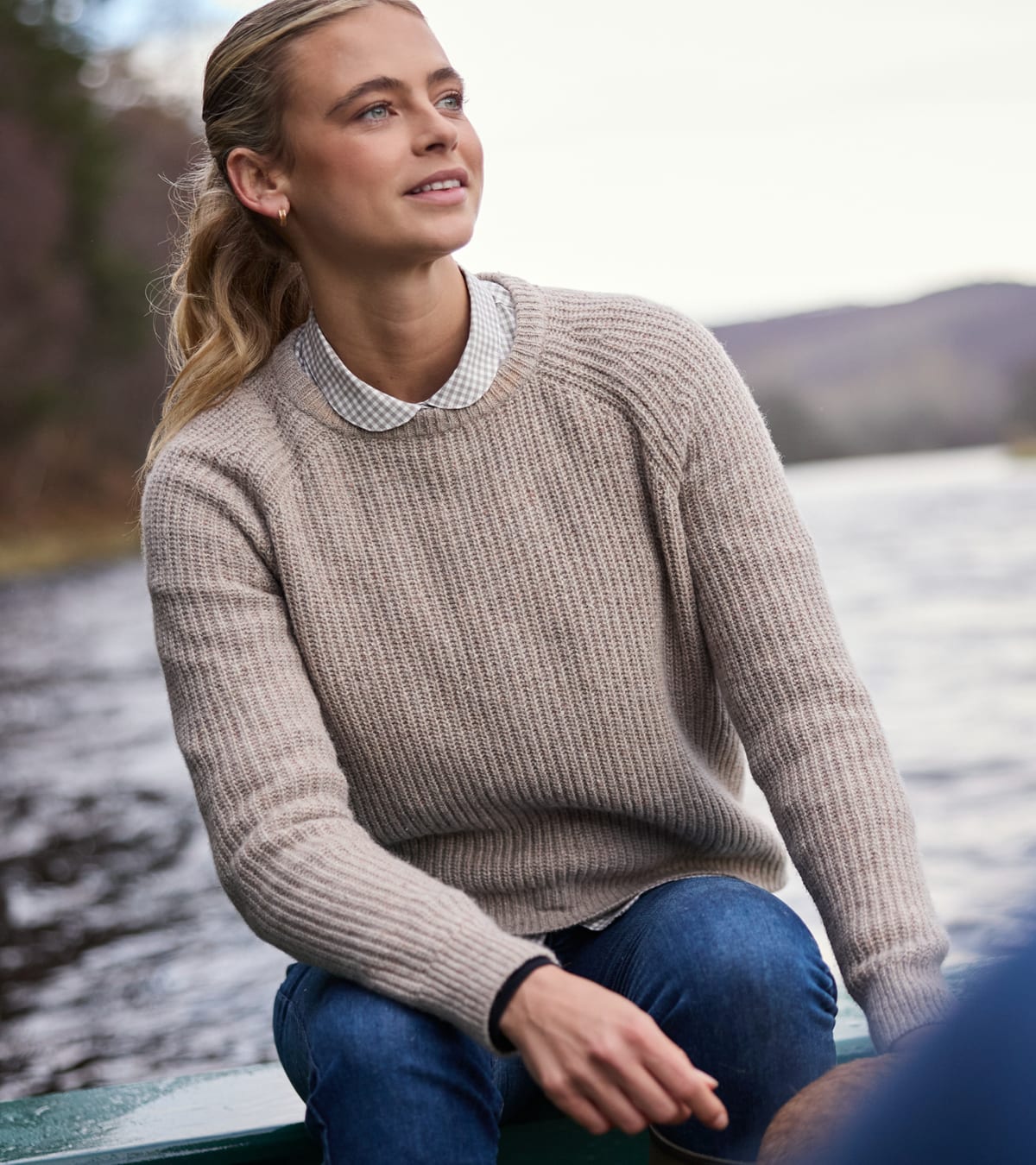 Woman wearing oatmeal ribbed knit jumper over gingham collared shirt, sitting in a boat by the water