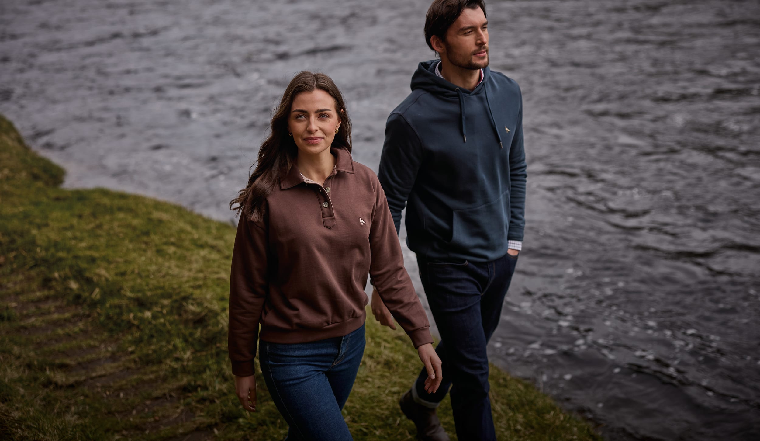 Man in navy quarter-zip pullover and woman in brown collared sweatshirt walking beside a riverbank
