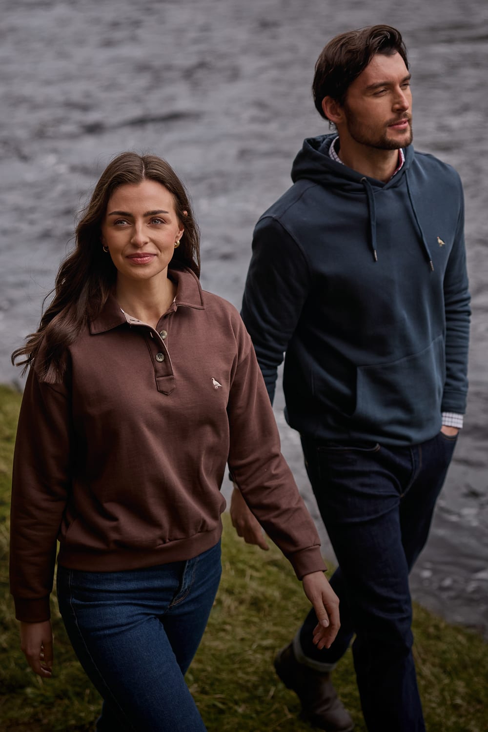 Woman and man walking by a river wearing brown polo sweatshirt and navy hoodie, mobile banner