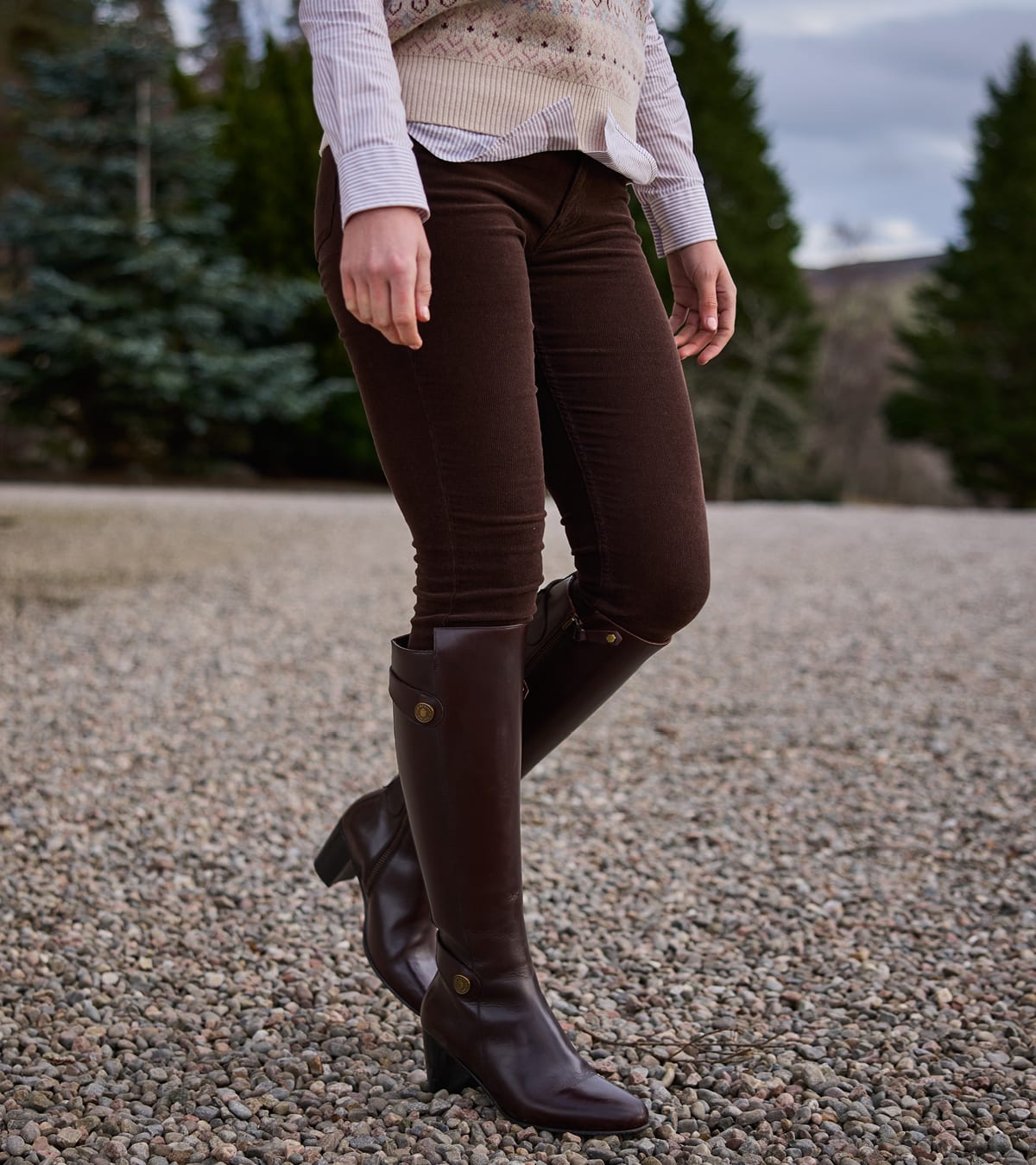 A woman outdoors wearing dark brown fitted trousers standing on a gravel path with trees in the background.