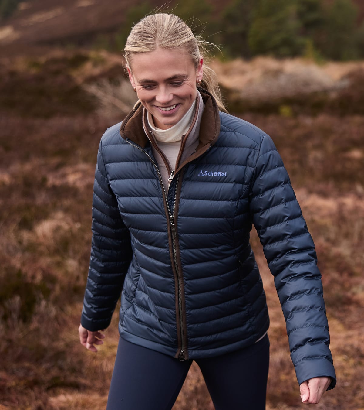 A woman wearing a navy blue insulated puffer jacket with a brown collar, walking outdoors in a grassy landscape.