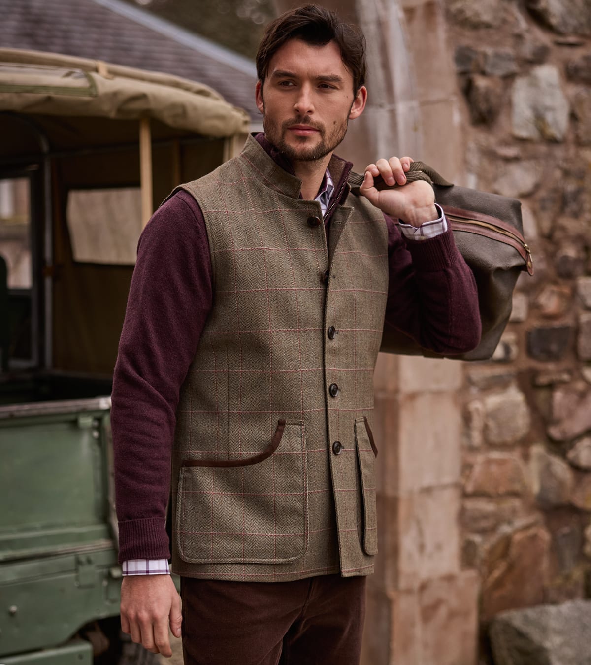 A man wearing a brown tweed waistcoat over a burgundy jumper, standing next to a stone wall.
