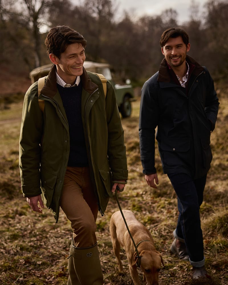 Two men walking outdoors with a dog on a leash, wearing outdoor jackets and boots.