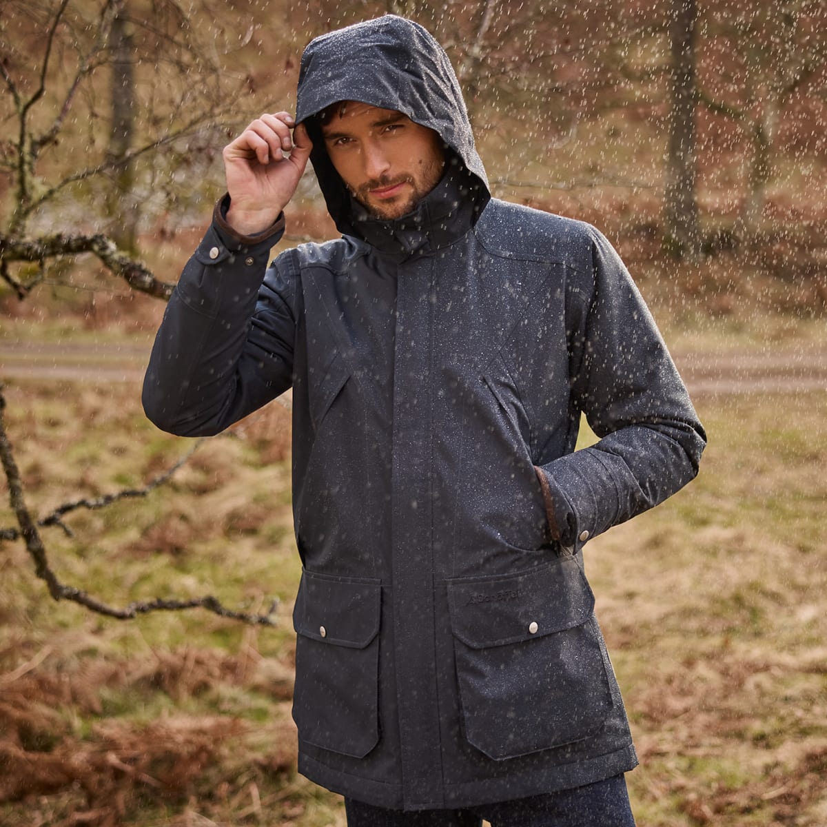 A man wearing a navy hooded rain jacket stands outdoors in falling rain, with a blurred forest background.