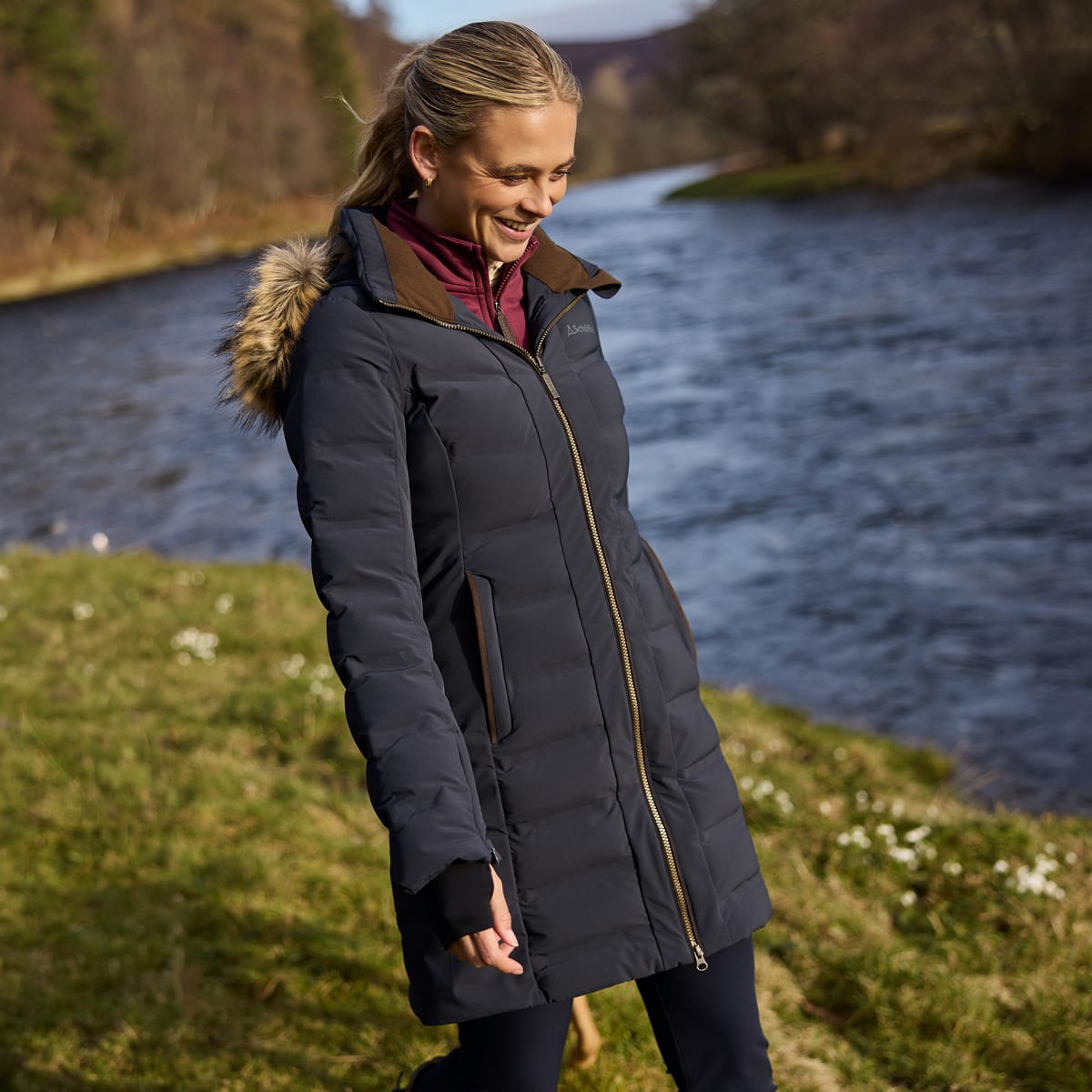 A woman wearing a navy puffer coat with brown trim and a faux-fur hood, standing outdoors next to a river.