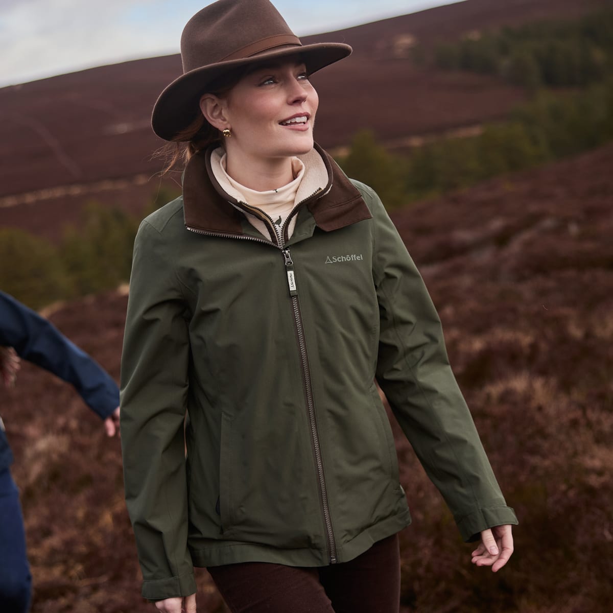 A woman wearing a green jacket with a brown collar standing in a moorland landscape.