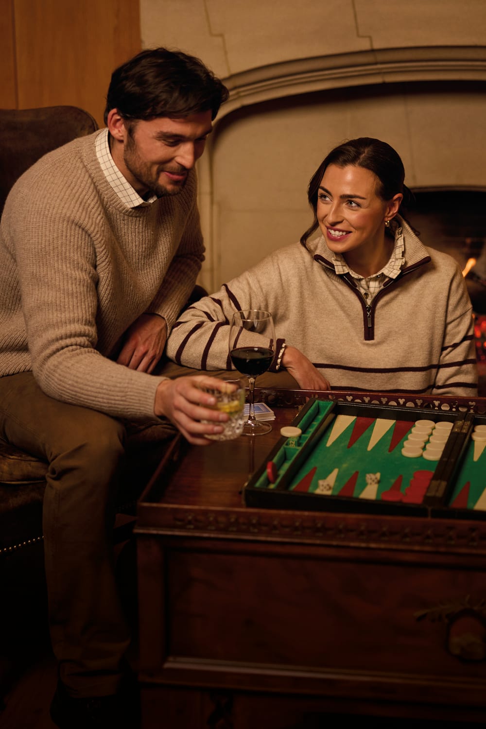 Man and woman playing backgammon by a fire in a cream knit jumper and striped quarter zip