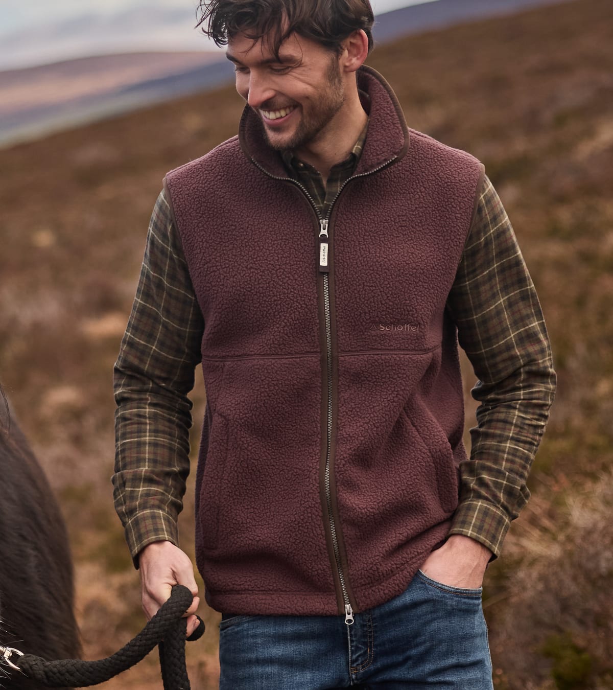 Man standing in moorland wearing a brown retro fleece holding onto a horse