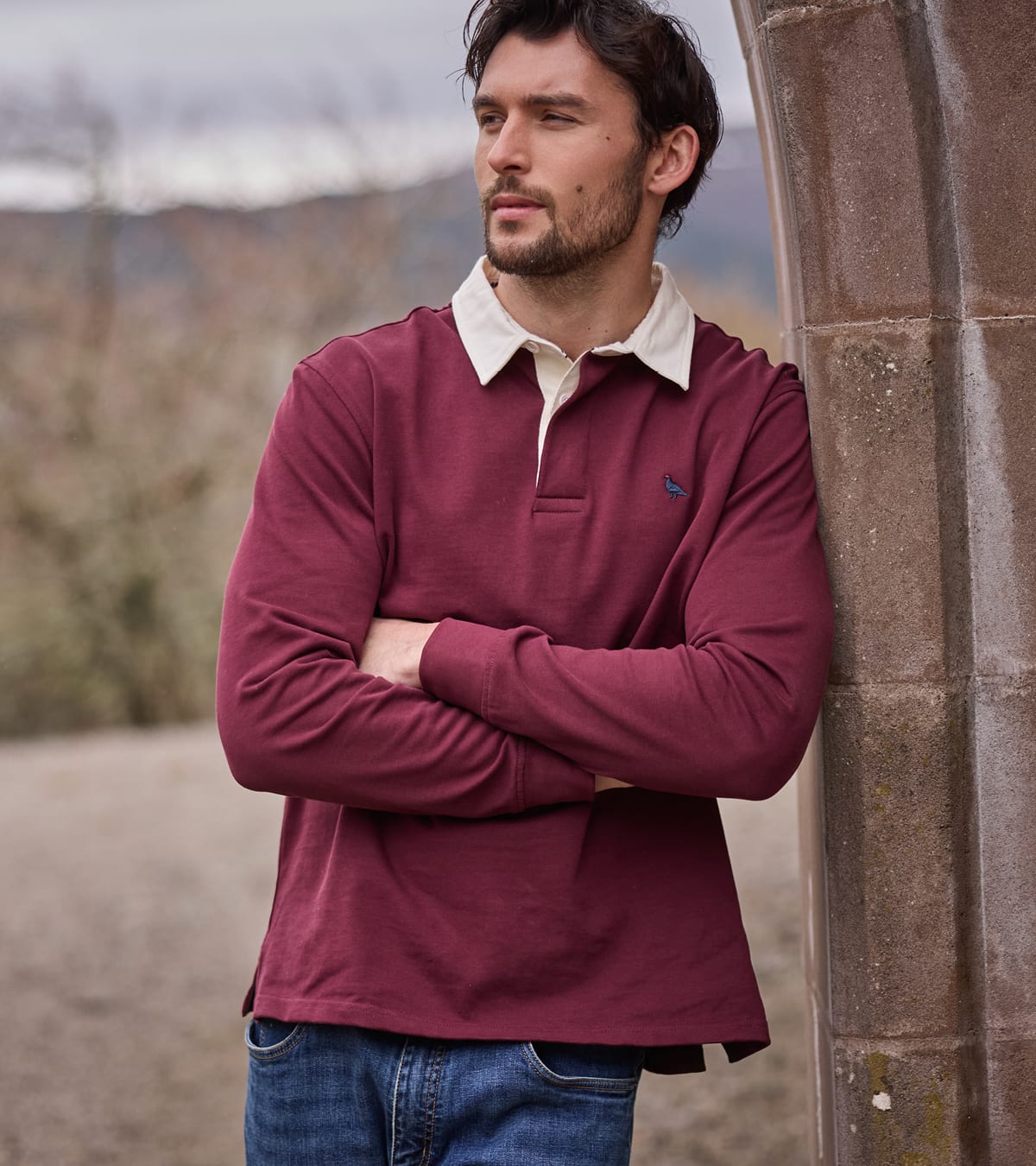 Man standing against a stone archway wearing a red rugby top and jeans