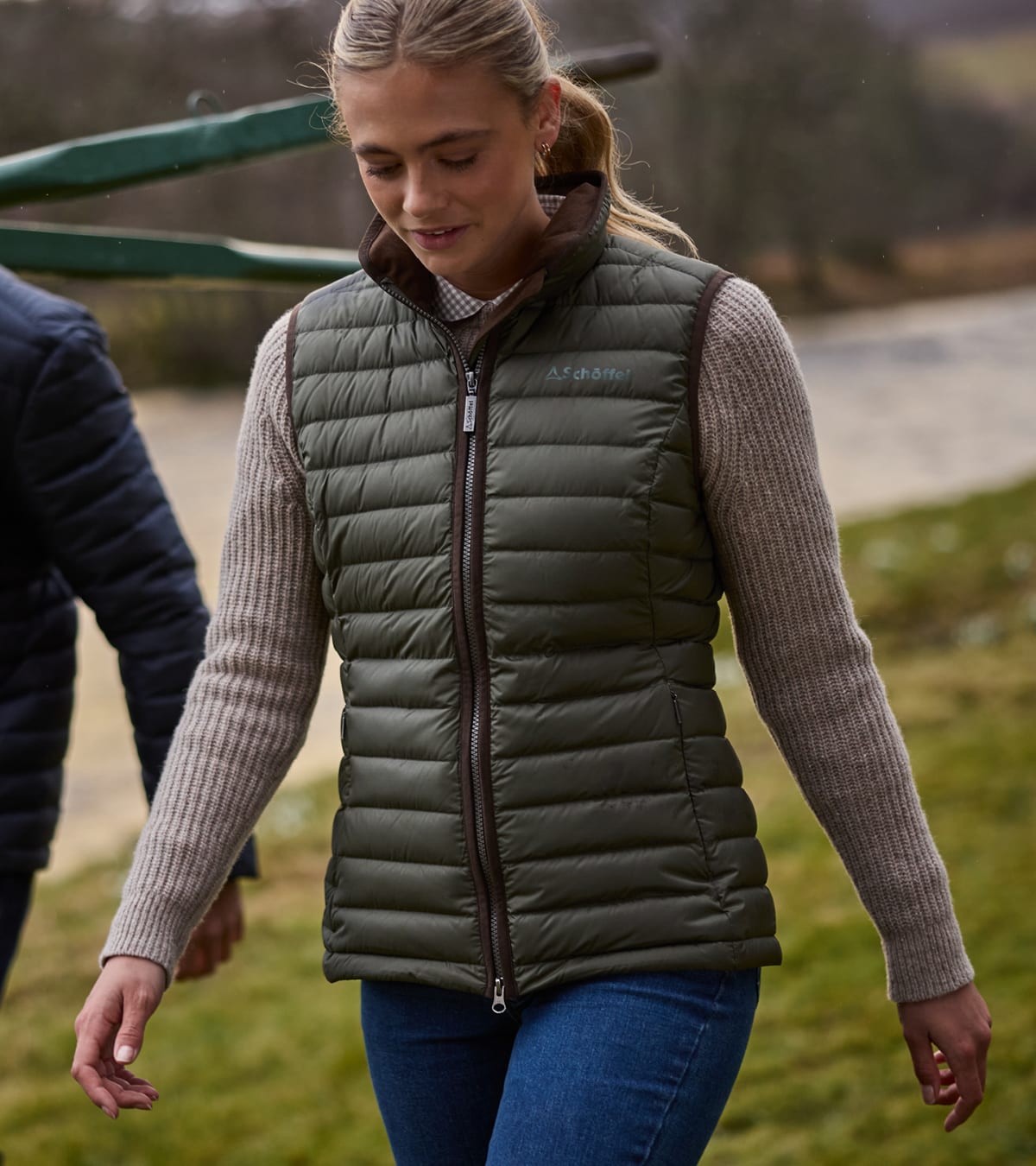 Woman walking near a body of water wearing a green padded gilet over an oat jumper.