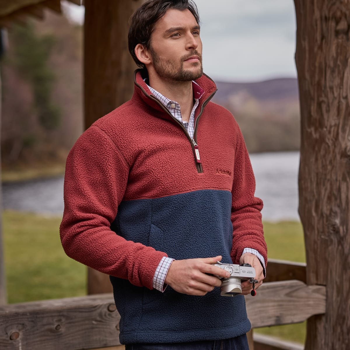 A man standing next to a log cabin wearing a two tone fleece pullover in red and blue holding a camera.