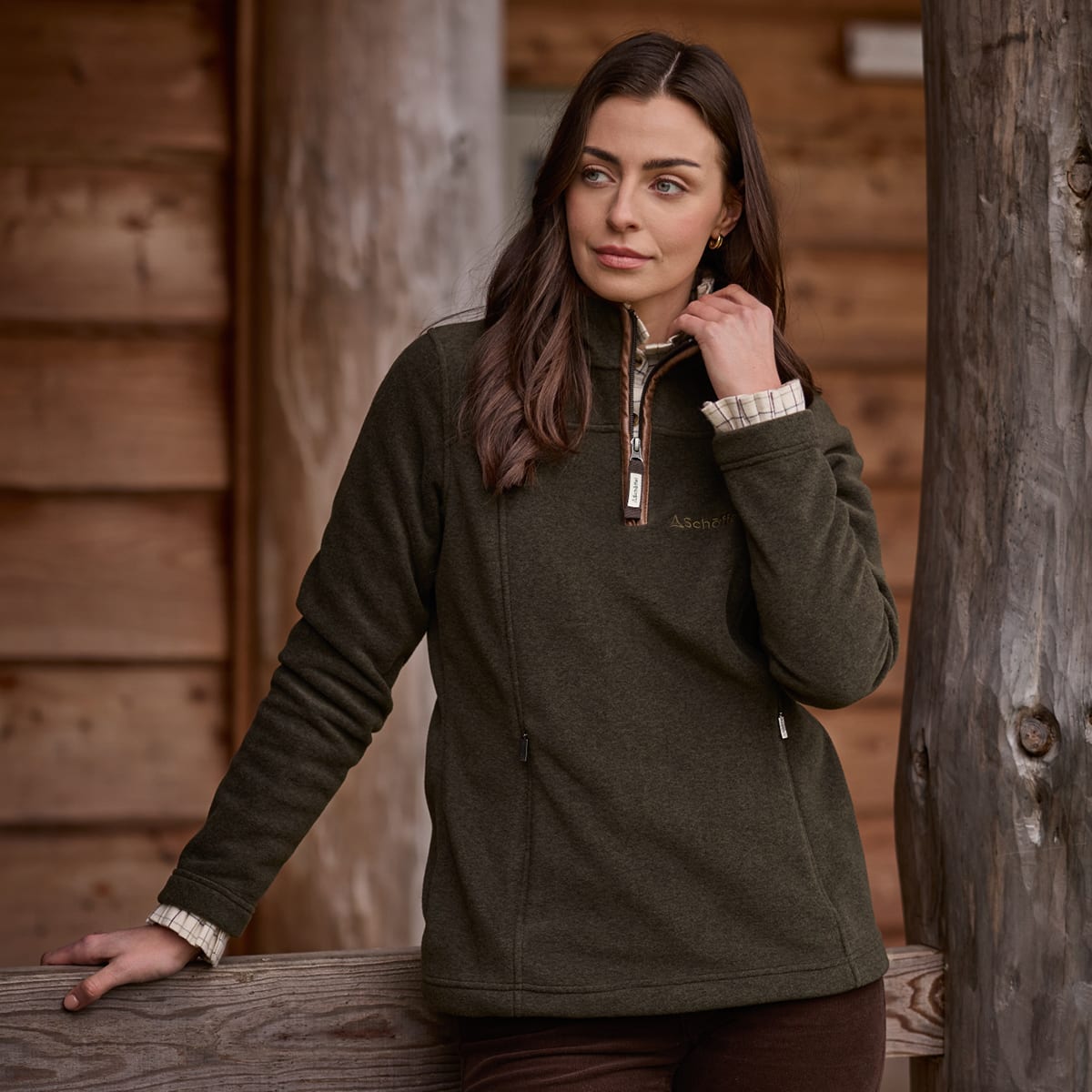 Woman standing near a log cabin wearing a green quarter zip fleece and brown trousers