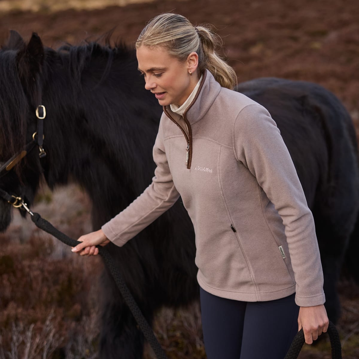 Woman leading a black horse across moorland wearing a beige quarter zip fleece