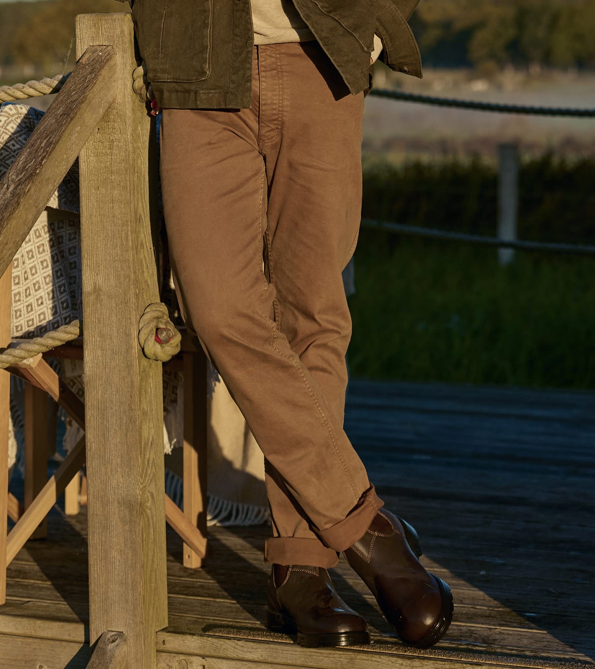 Person wearing brown cuffed trousers, dark boots and a jacket on a wooden deck surrounded by greenery.