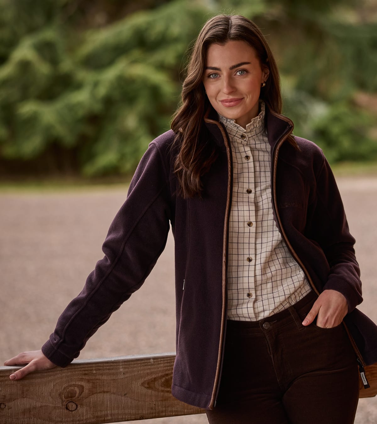 Woman wearing a plum fleece jacket, checked shirt and brown trousers, leaning on a wooden fence outdoors.