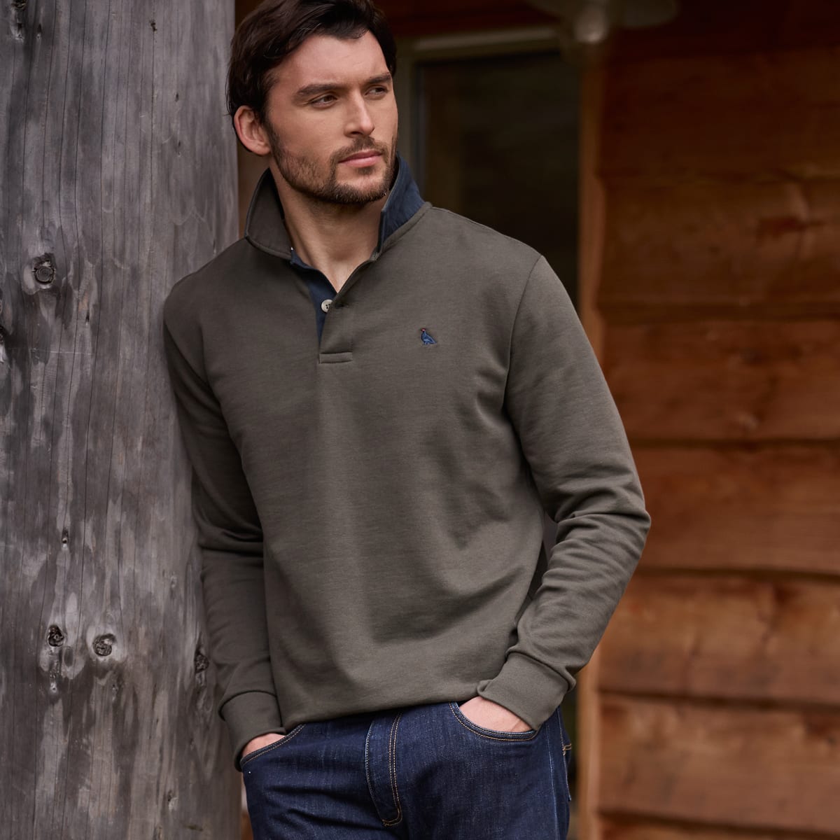 Man wearing an olive green quarter-zip pullover with a navy collar, leaning against a wooden post.
