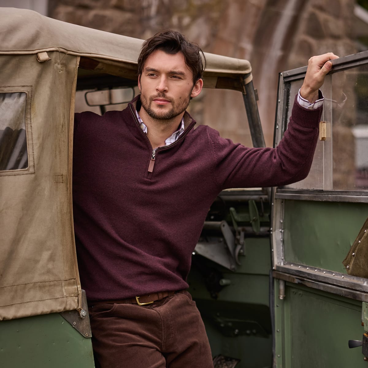 Man wearing a burgundy quarter-zip sweater and brown corduroy trousers beside a vintage green vehicle.
