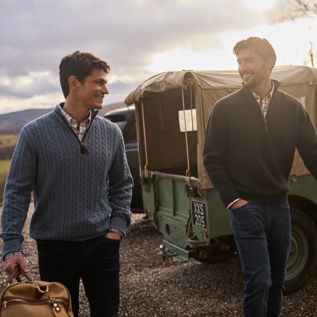 Two men in quarter-zip sweaters standing beside a vintage green jeep, one holding a tan bag.