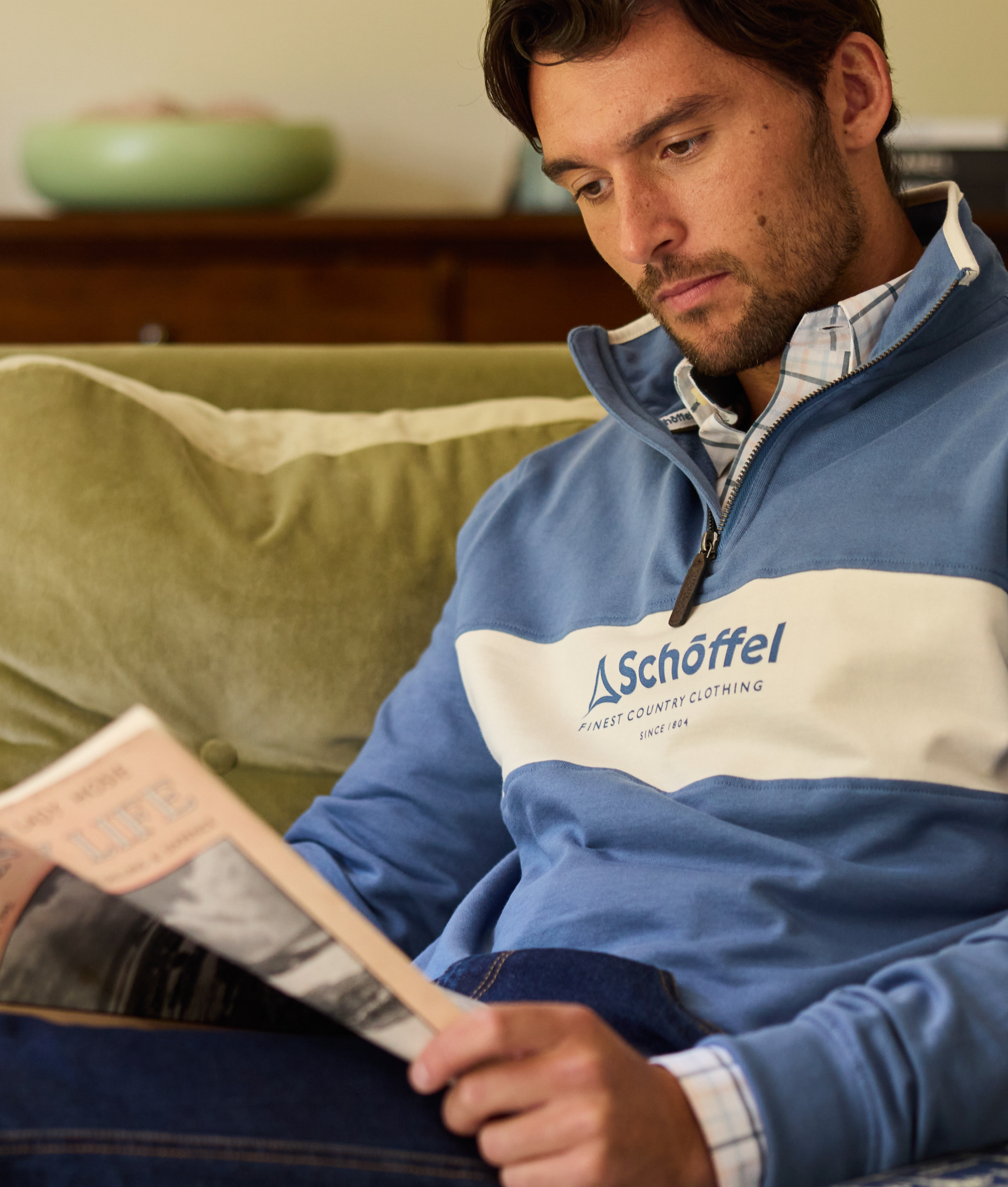 Man wearing a blue and white jacket with 'Schoffel' logo, reading a magazine on a couch.