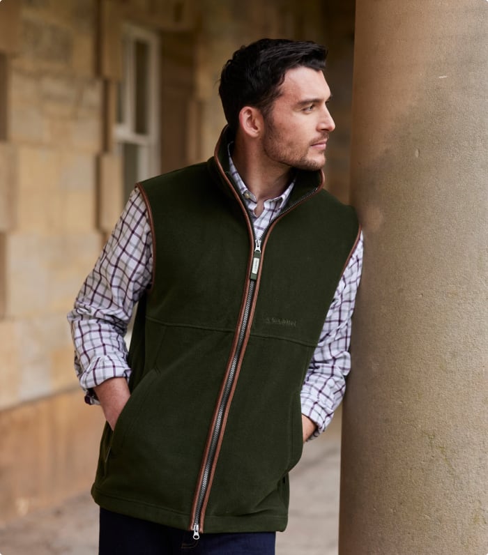 A man leans against a stone pillar wearing an olive green fleece gilet with tan zip trim over a checked shirt, gazing into the distance.