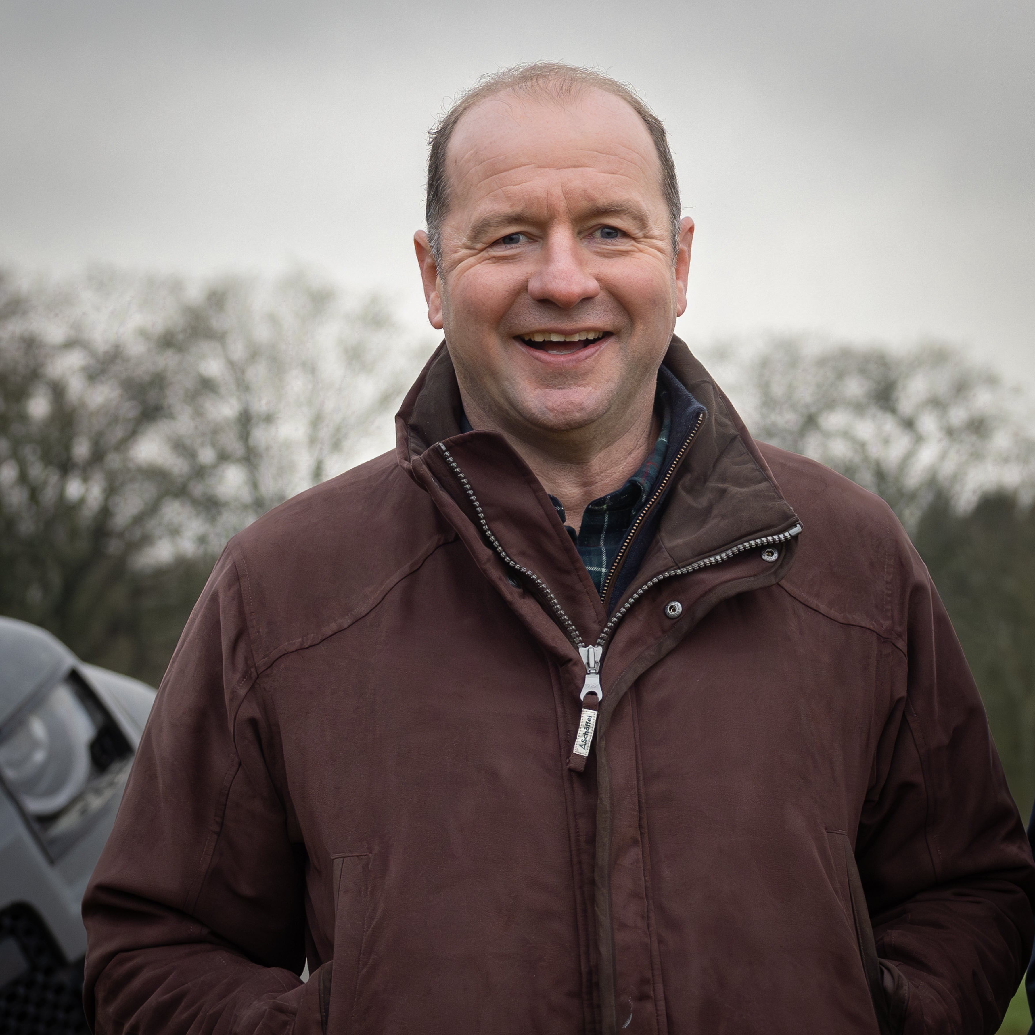 Geordan Murphy wearing a brown jacket standing in a country setting with trees in the background