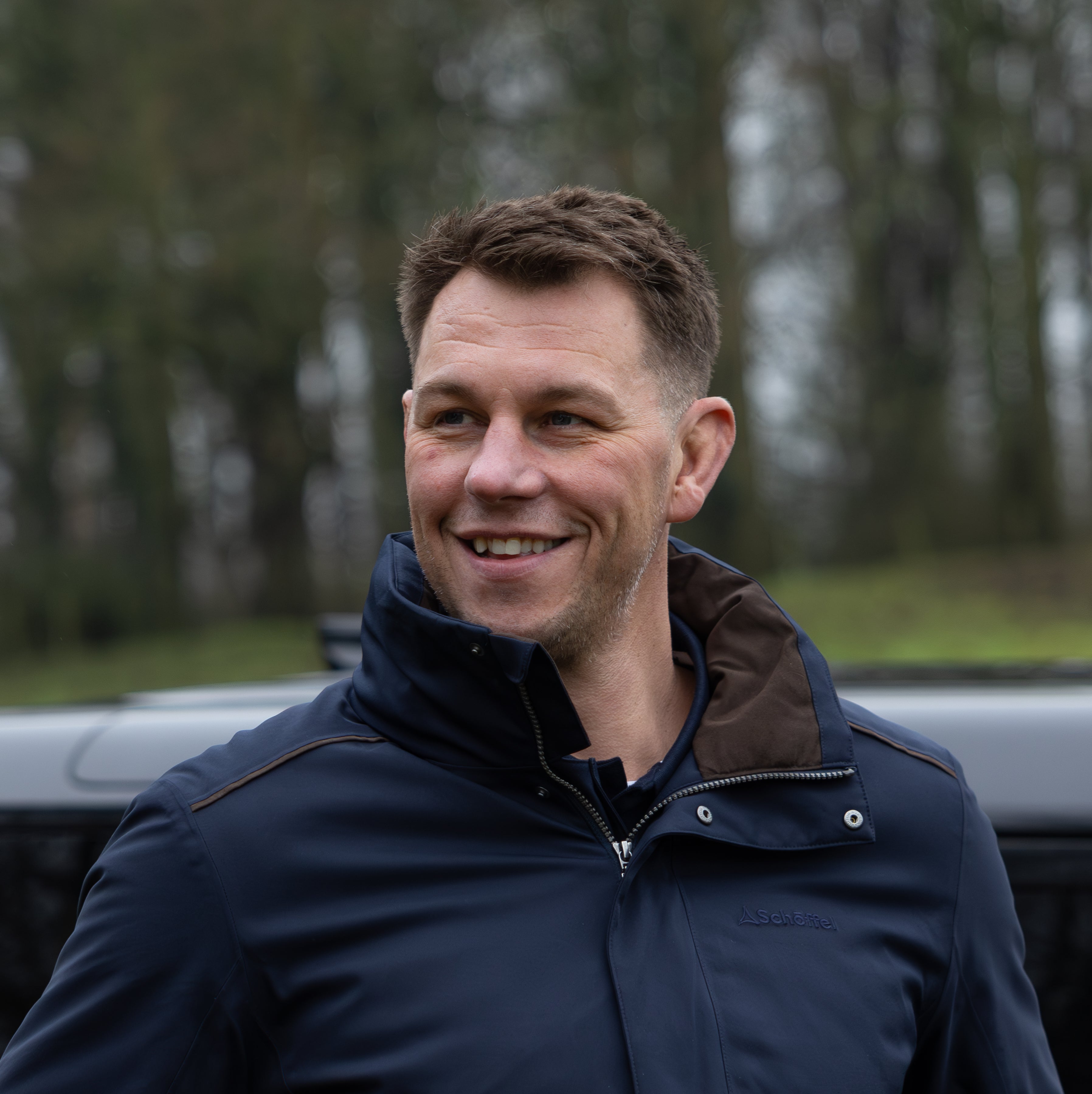 Louis Decan wearing a blue jacket.  He is standing in front of a car in a woodland setting.