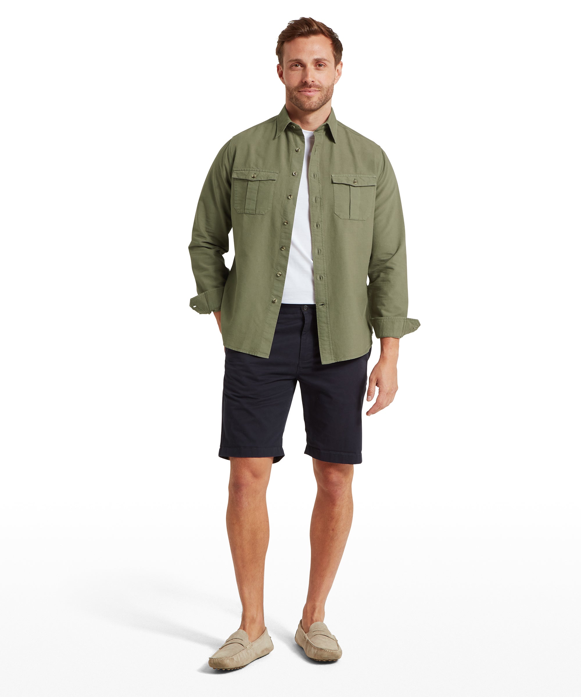 A man dressed in a light olive green button-up shirt with long sleeves and two chest pockets, worn open over a white T-shirt. The sleeves are rolled up slightly. He pairs the shirt with dark navy shorts and light beige loafers, creating a relaxed, warm-weather outfit.