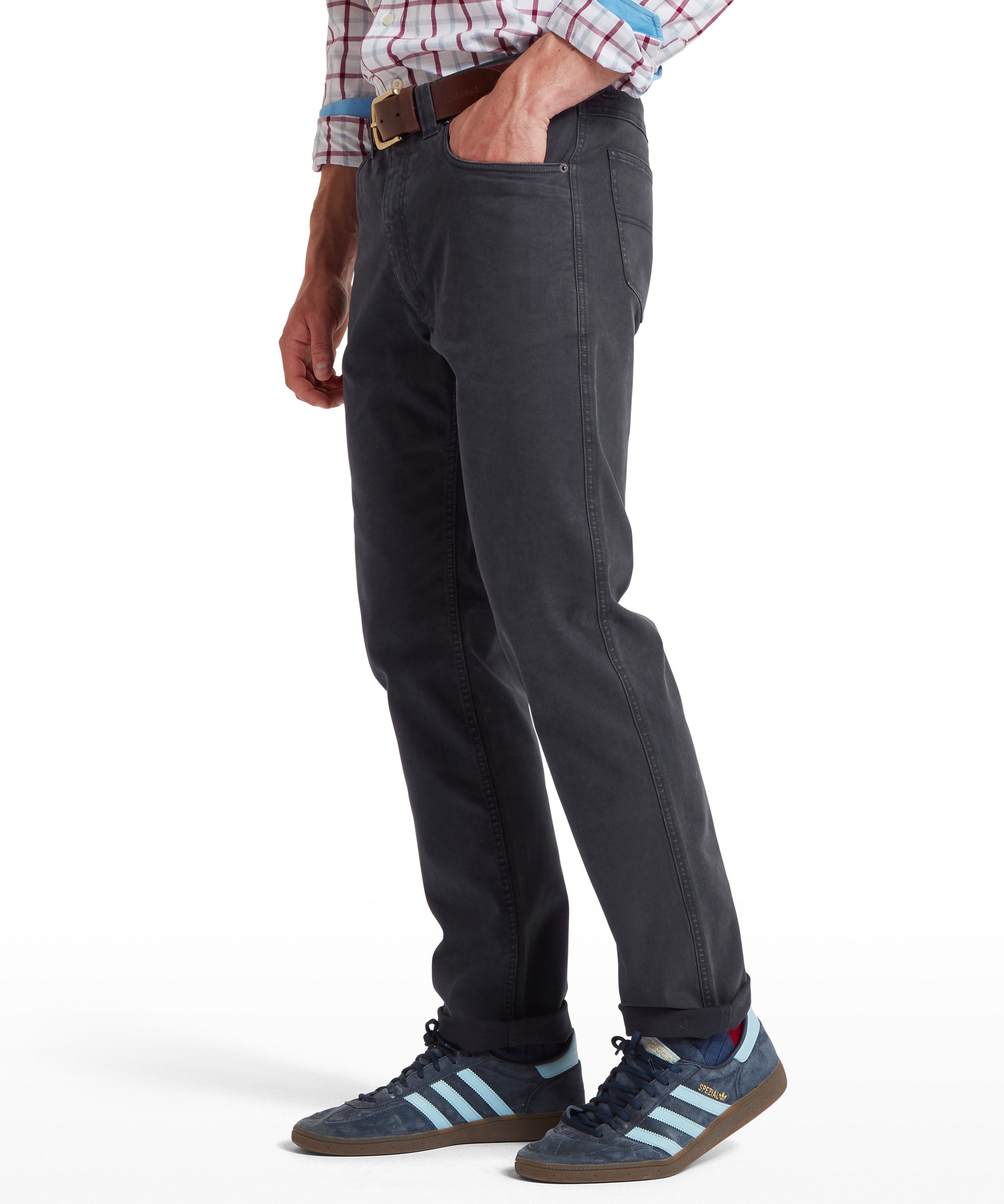 Lower half view of a man wearing dark grey trousers with a straight fit, brown belt and dark blue trainers with light blue stripes and gum soles. Trousers are cuffed at the ankles; one hand is in a pocket. Outfit shown on a transparent background