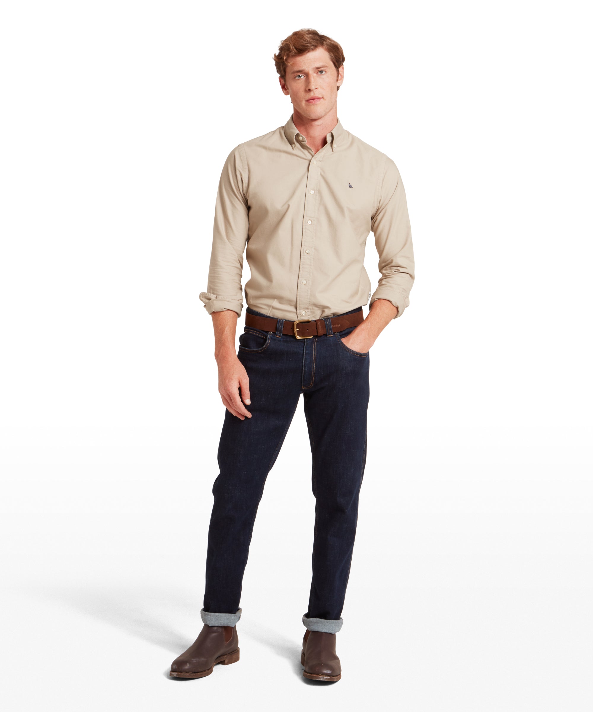 Man wearing a beige shirt, dark jeans, and brown shoes on a white background