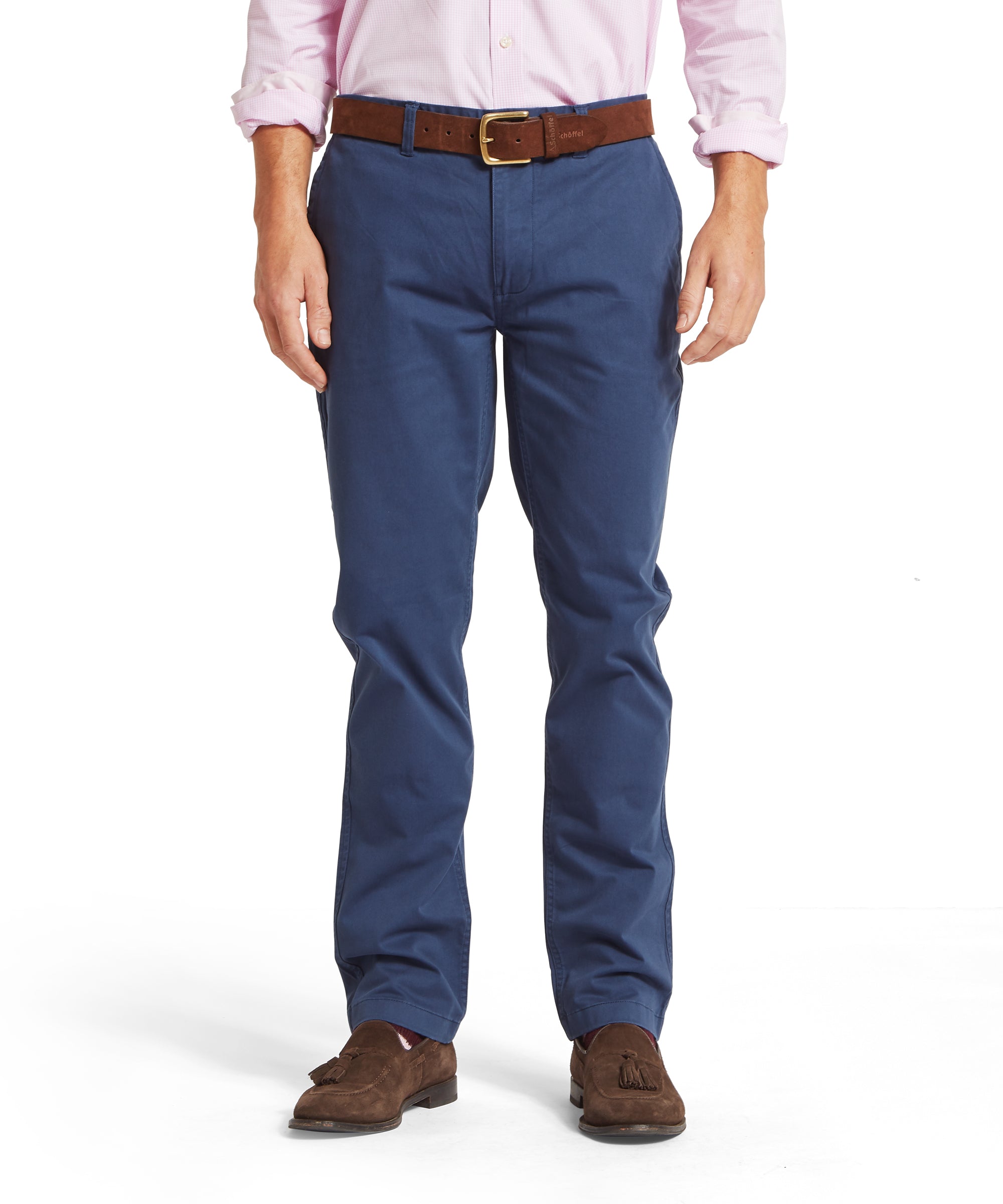 Man wearing a pale pink check shirt with rolled-up sleeves, tucked into blue chinos, styled with a brown belt with gold buckle and brown suede tassel loafers. Outfit shown in a relaxed standing pose on a plain white background.