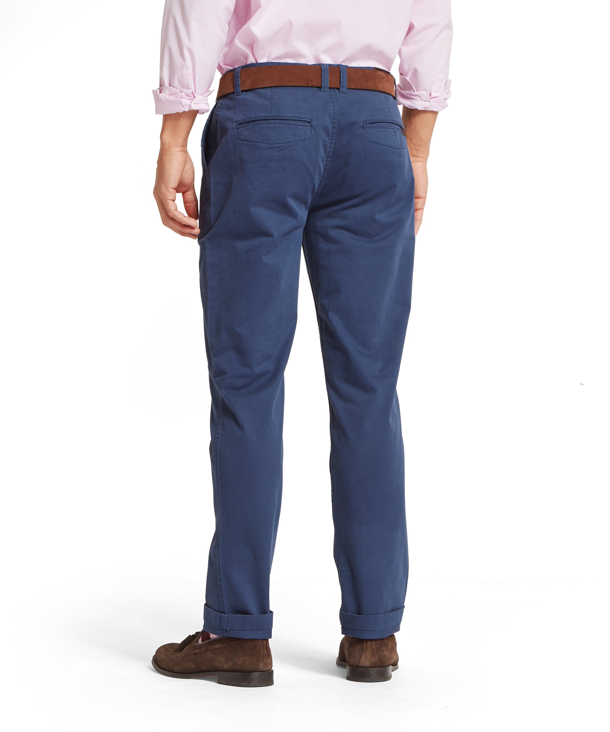 Rear view of a man wearing a pale pink shirt with rolled-up sleeves, tucked into blue chinos with back welt pockets, styled with a brown belt and brown suede shoes. Chinos are cuffed at the ankles. Pose shown on a plain white background
