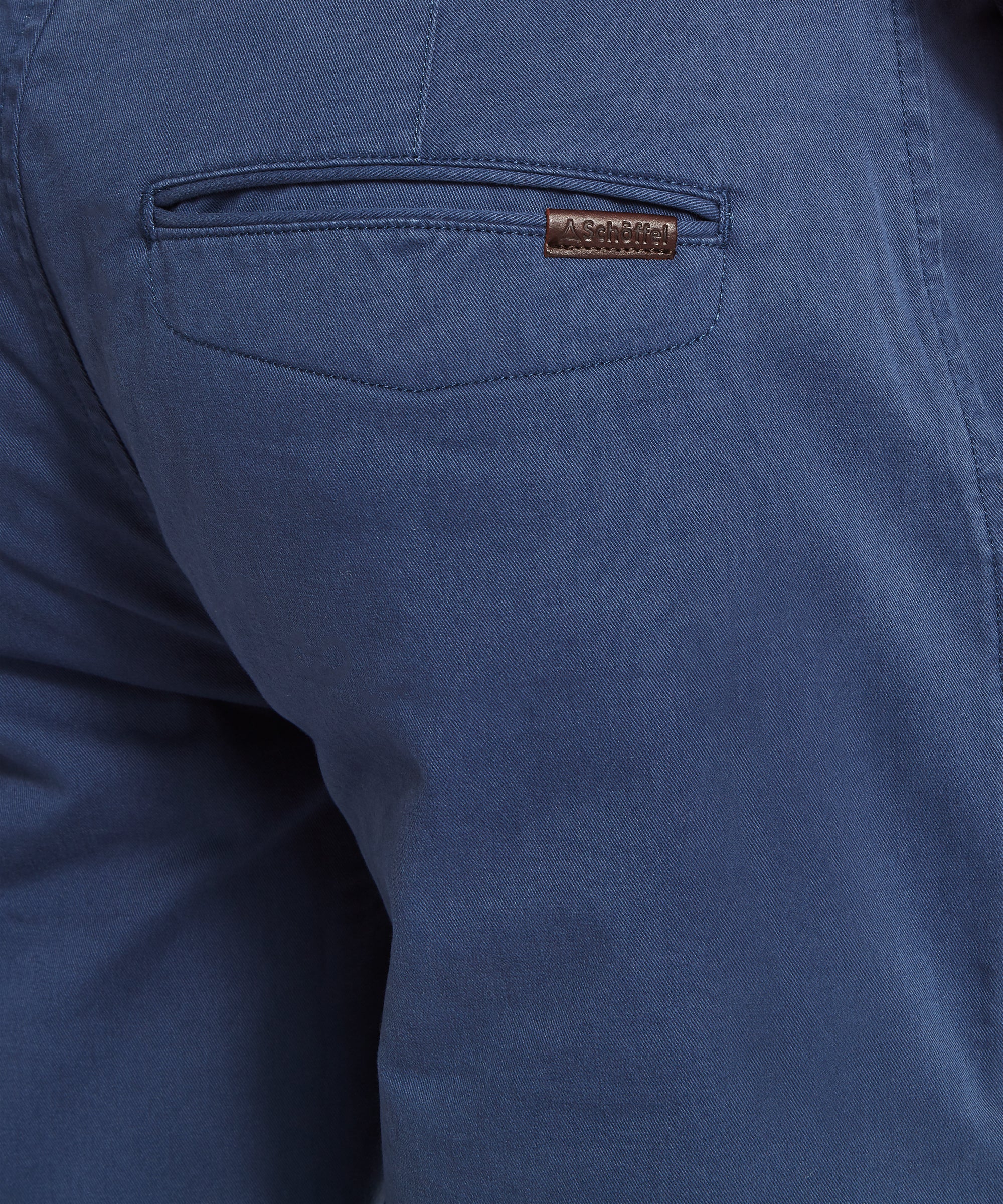 Close-up rear view of blue chinos with welt pocket and small Schöffel brand tag above the pocket. A braided blue belt is threaded through the loops. Shown on a plain white background
