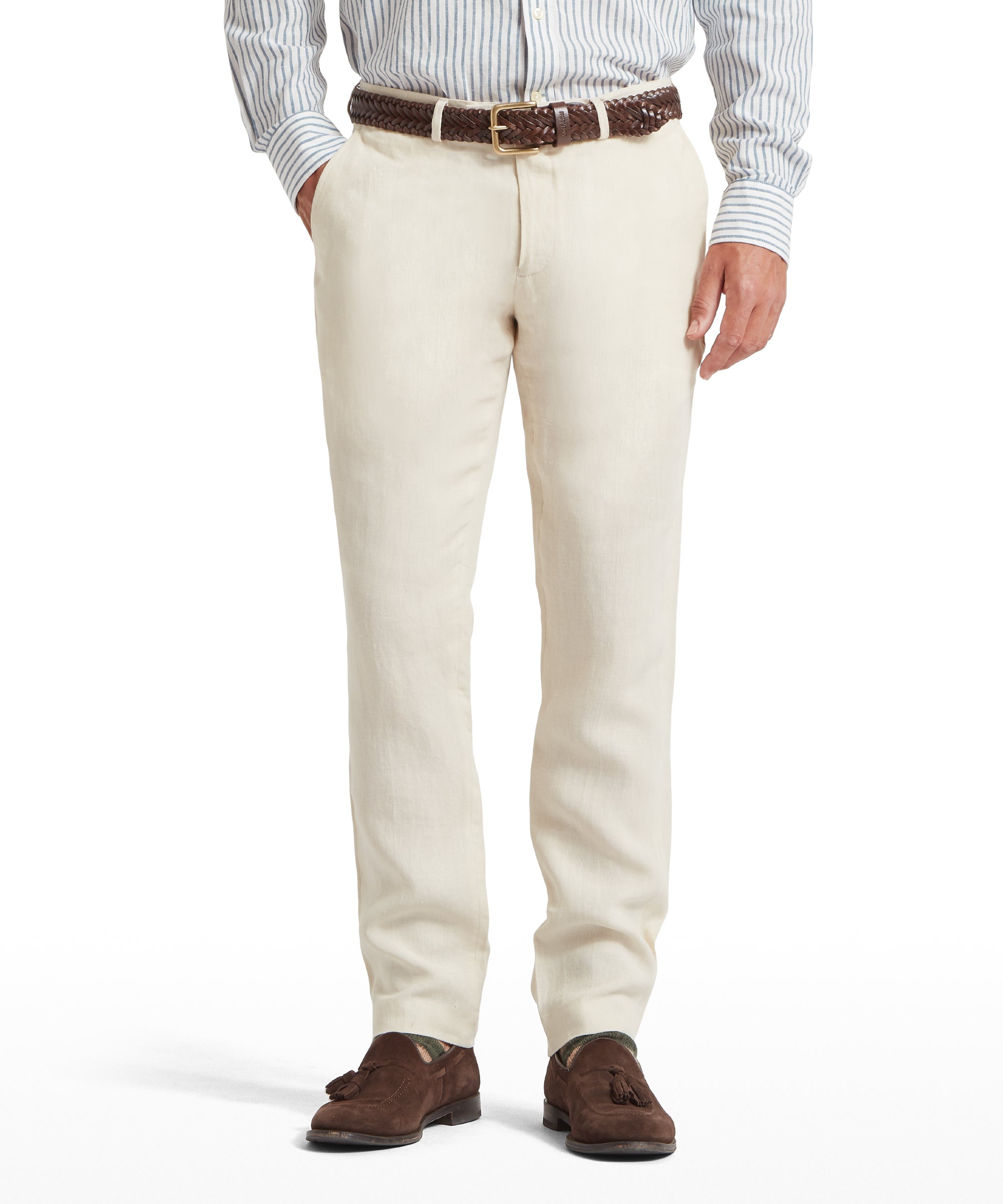 Lower half of a man wearing cream linen trousers with a brown woven belt featuring a gold buckle. Paired with a blue and white striped button-down shirt and brown suede tassel loafers. Standing with hands in pockets against a plain white background.