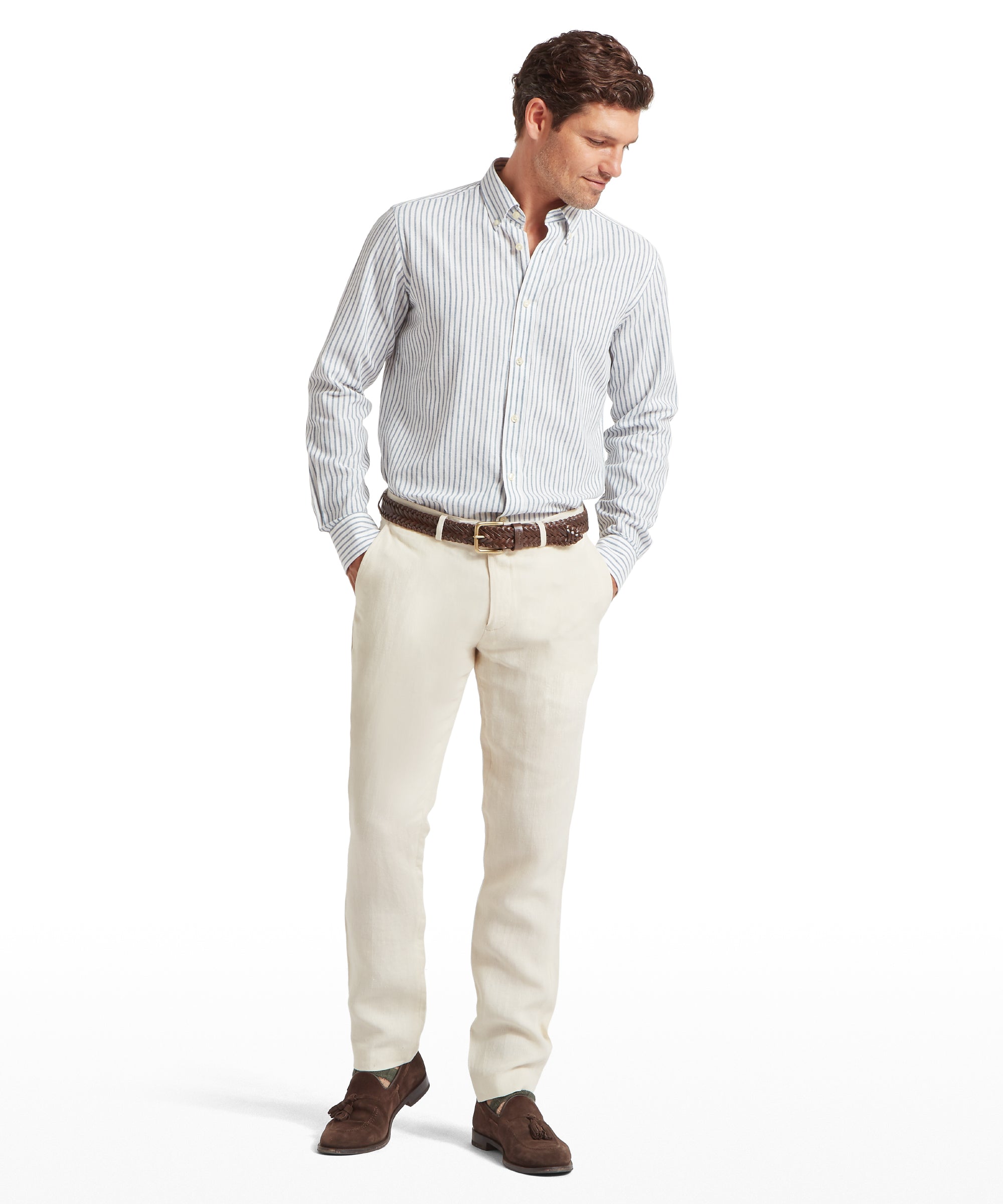 Man wearing a light blue and white vertically striped button-down shirt tucked into cream linen trousers. Styled with a brown woven belt and brown suede loafers. Looking slightly downward against a plain white background.
