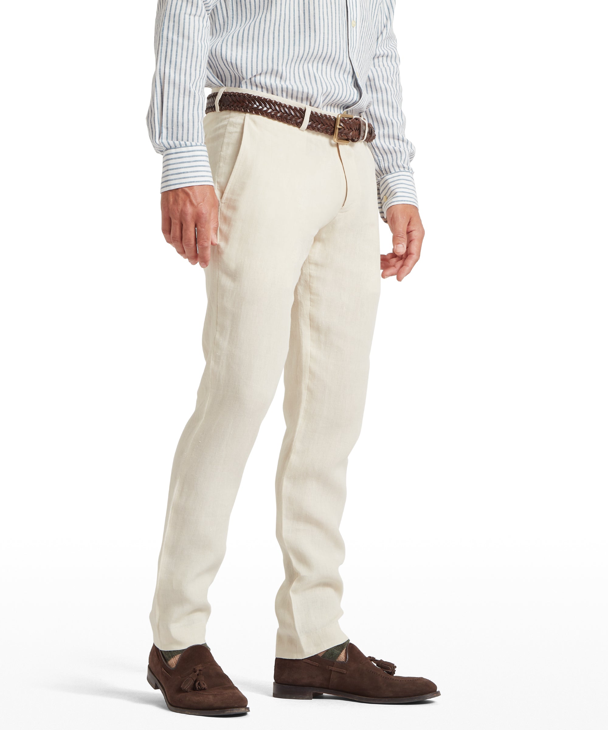 Man wearing cream linen trousers with a tailored fit and clean front. Paired with a light blue and white striped long-sleeved shirt, a brown woven leather belt, and brown suede tassel loafers. Standing front-facing against a plain white background.