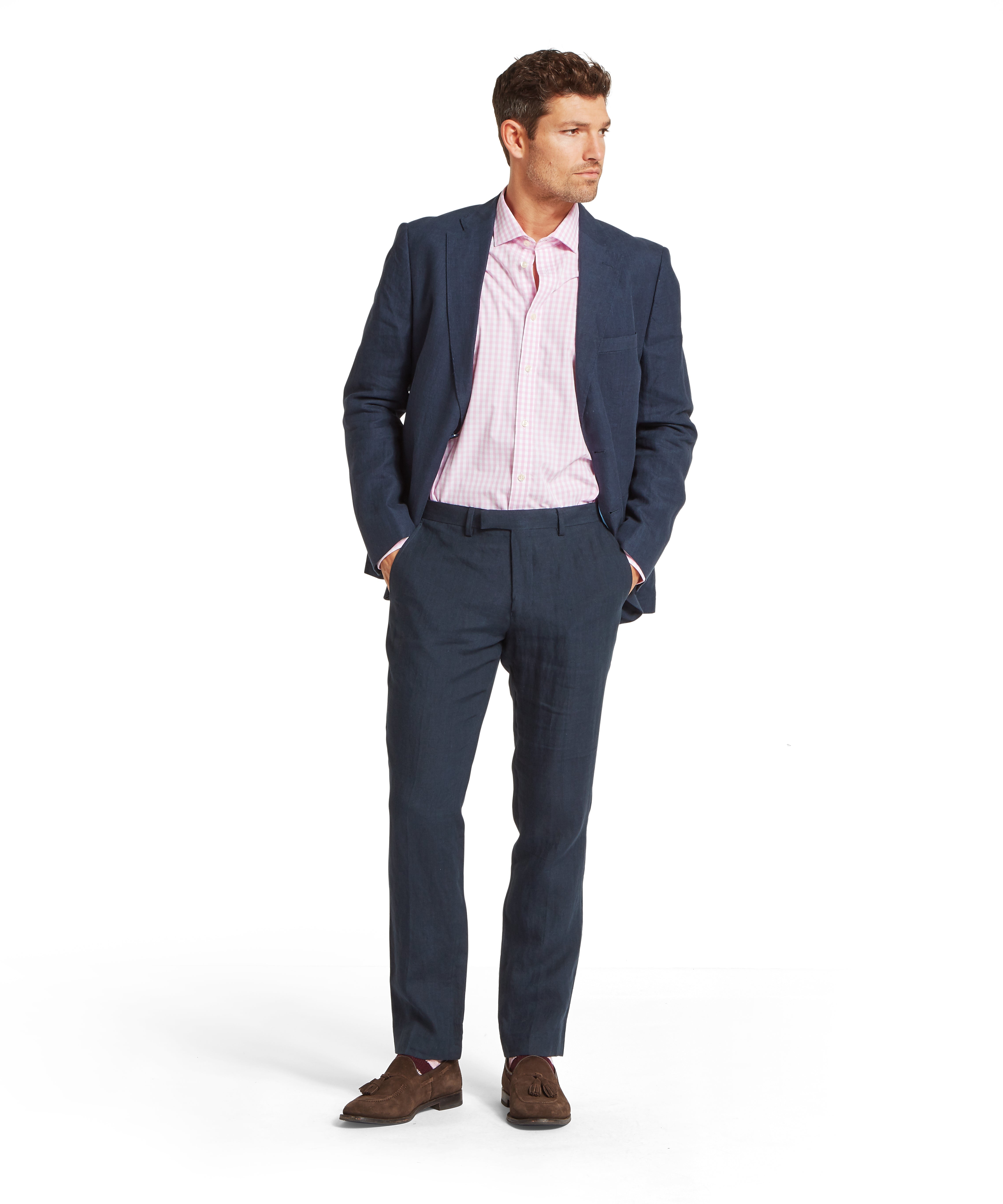 Man wearing a dark blue suit with matching blazer and trousers, paired with a light pink checked shirt. Styled with brown tassel loafers. Standing with hands in pockets against a plain white background.