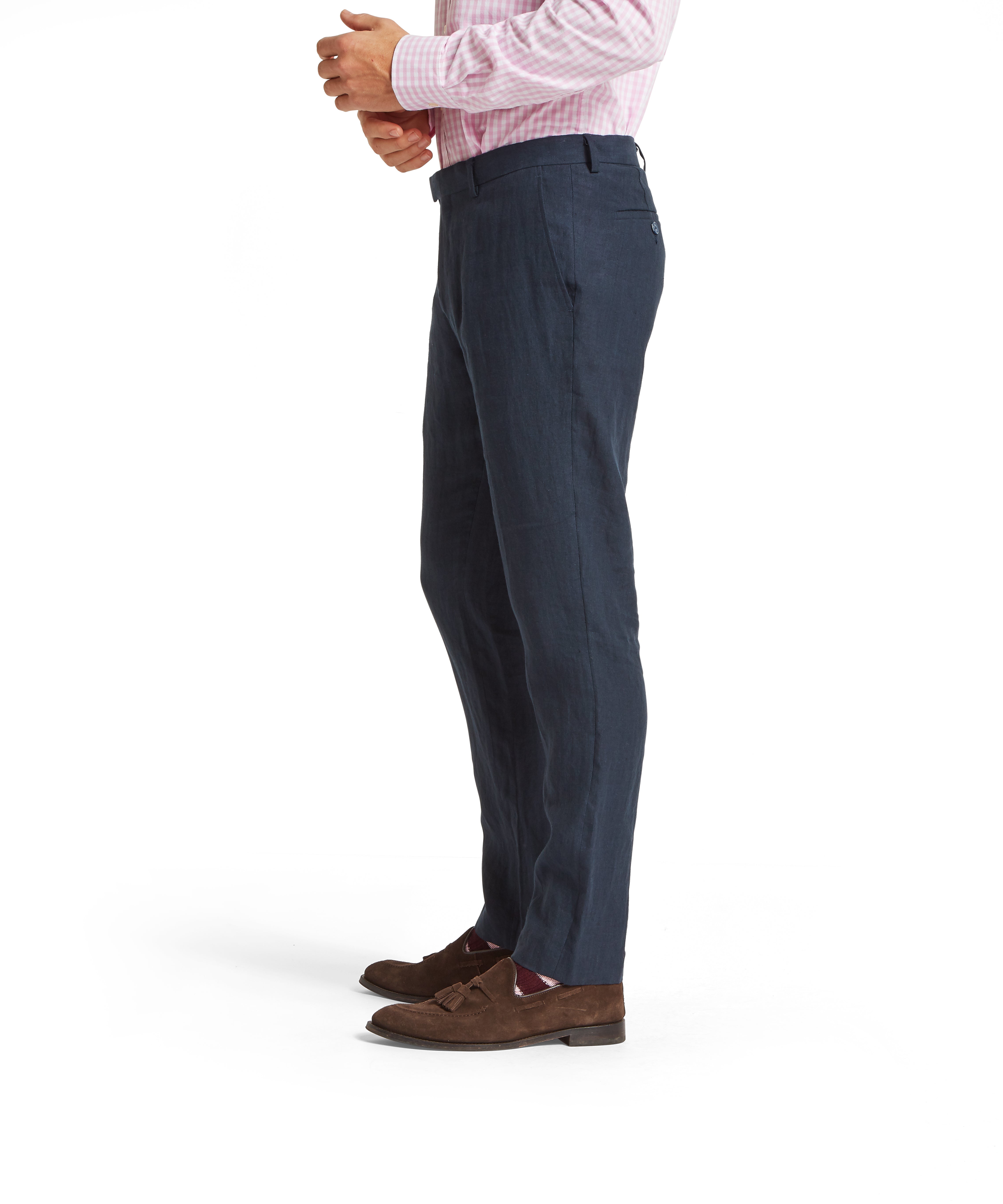 Side profile of a man wearing a light pink and white check shirt tucked into navy linen trousers. The trousers have a tailored fit with front creases and belt loops. Styled with brown suede tassel loafers and red patterned socks with white accents. Standing against a plain white background.