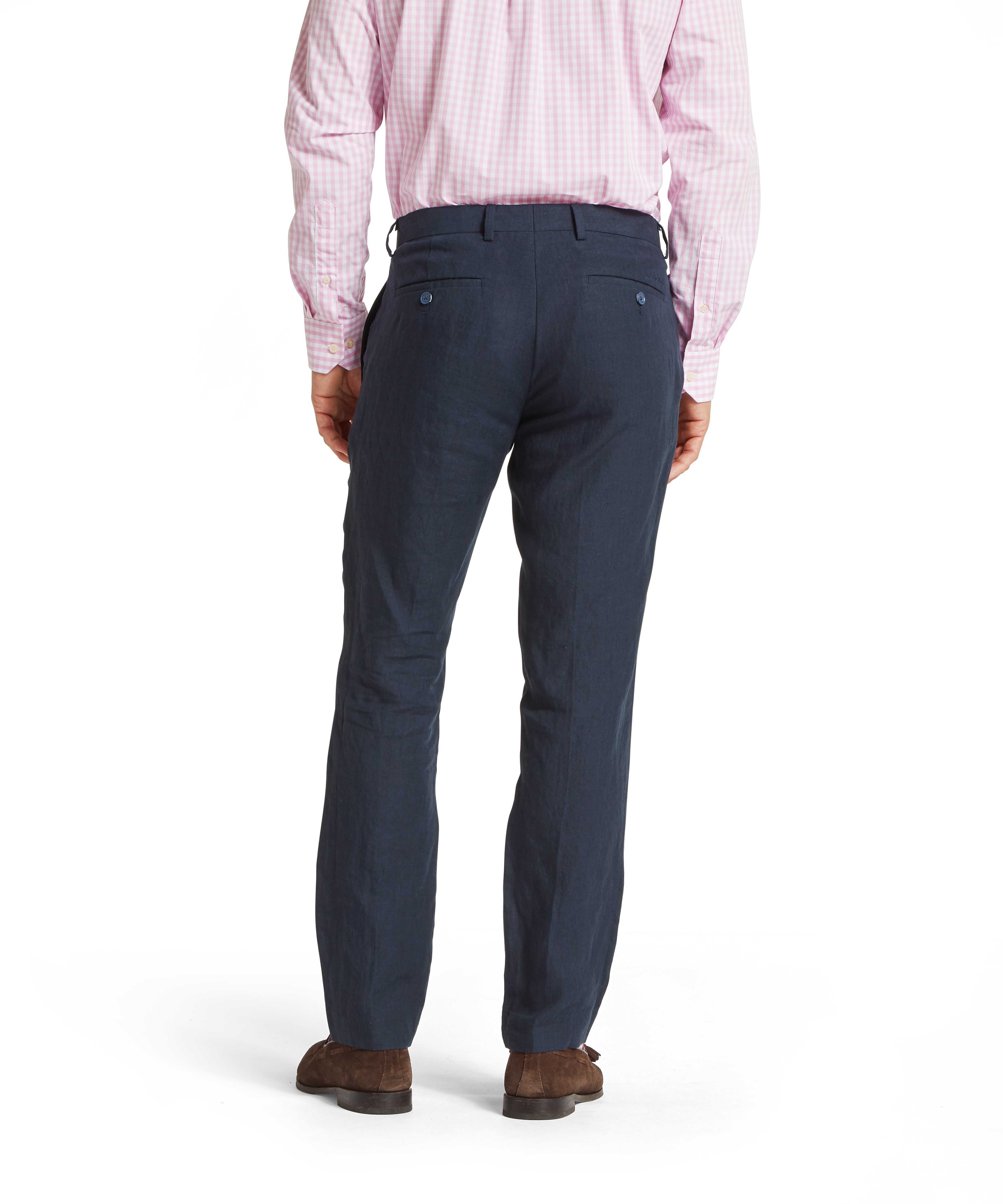 Back view of a man wearing a light pink check long-sleeved shirt tucked into navy linen trousers. The trousers feature two buttoned back pockets. Styled with brown suede shoes. Standing upright against a plain white background.
