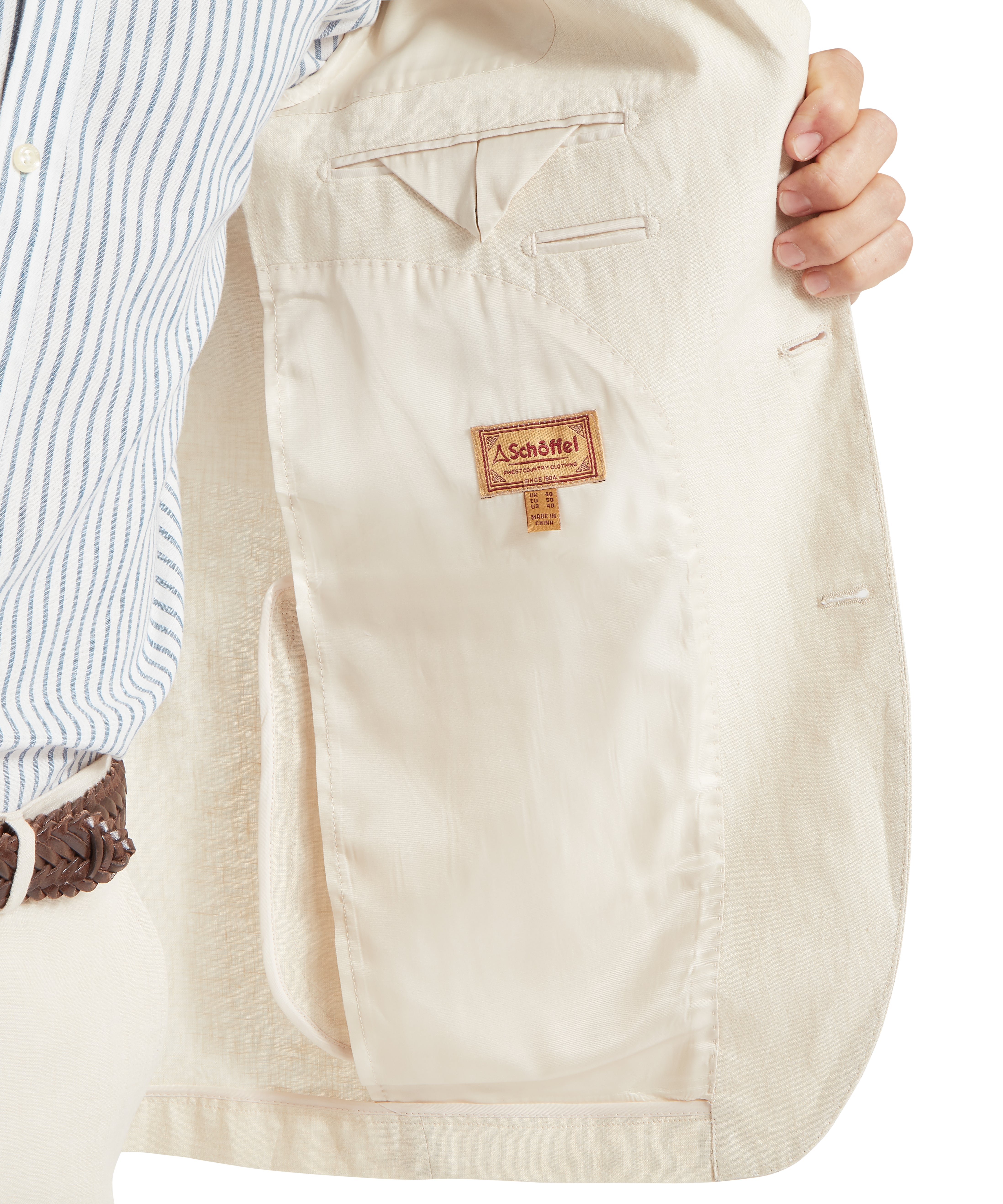 Man holding open a cream jacket to reveal its interior. The lining is light-coloured with a visible label reading “ Schöffel.” The inside features two horizontal slit pockets and a larger vertical pocket on the lower left side. He is wearing a blue and white striped shirt and a brown woven belt. The image highlights the craftsmanship and interior detailing of the jacket.