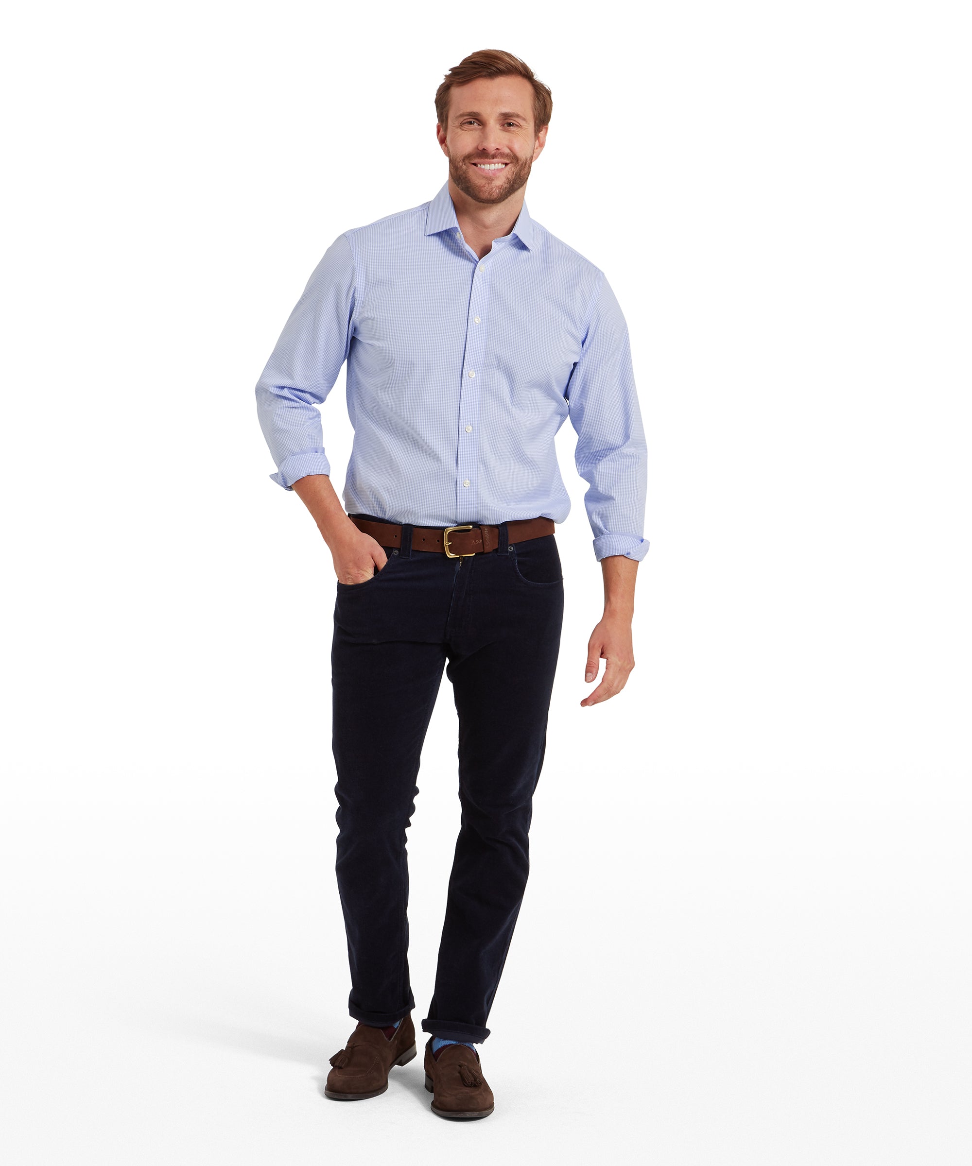 Man wearing a light blue, long-sleeved, button-down shirt with a subtle check pattern, paired with dark navy trousers, a brown belt, and brown suede shoes. One hand is in his pocket while the other arm hangs naturally. He has short hair and a neatly groomed beard. The shirt sleeves are rolled up slightly. He is standing against a plain white background. The outfit is casual yet polished, suitable for various occasions.
