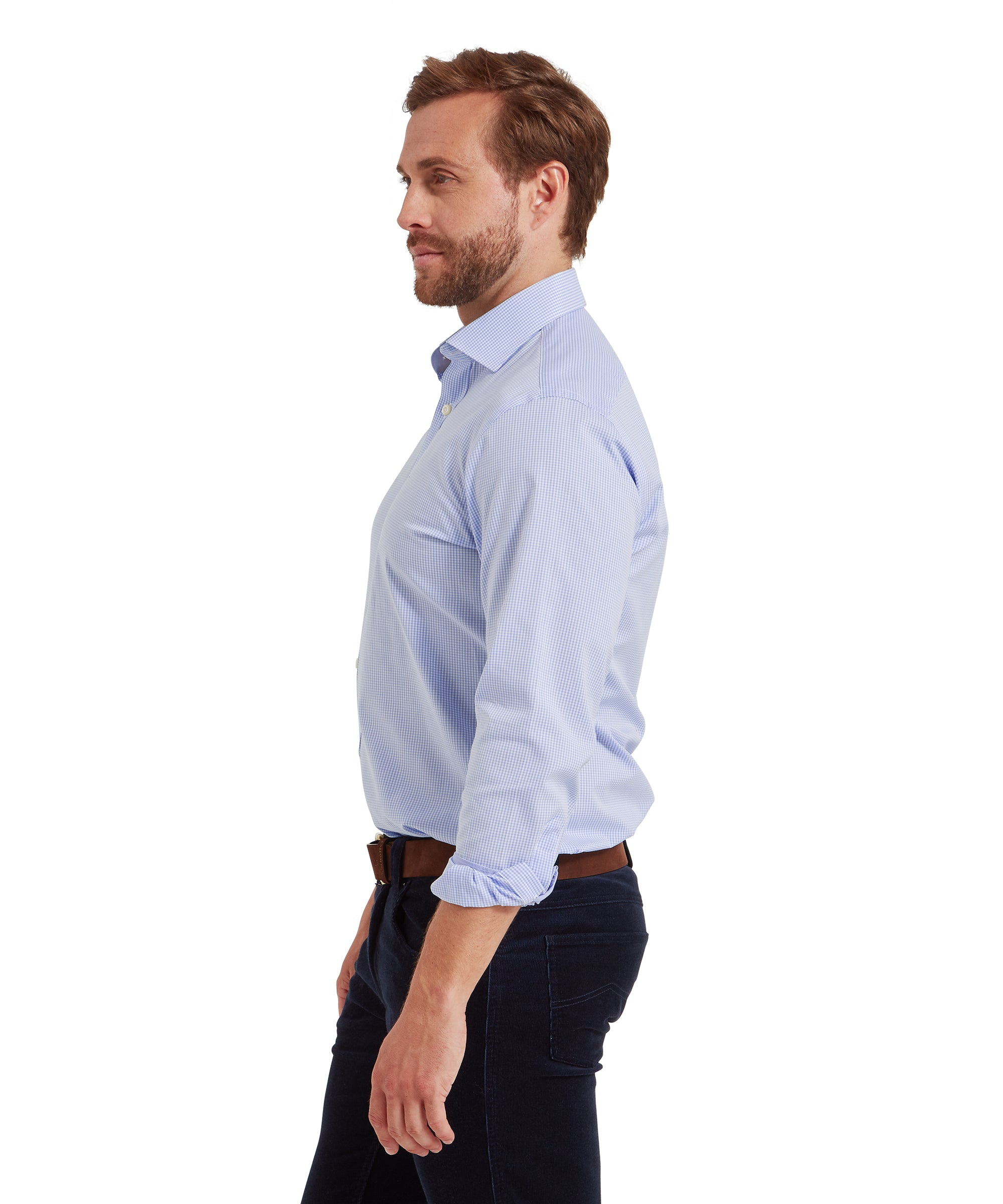 Man standing in profile view, facing left, wearing a light blue, long-sleeved, button-up shirt with a subtle vertical stripe pattern. The shirt is tucked into dark trousers and secured with a brown belt. The sleeves are rolled up to the forearms. He has short, neatly styled hair and a trimmed beard. The plain white background keeps the focus on the fit and style of the shirt.