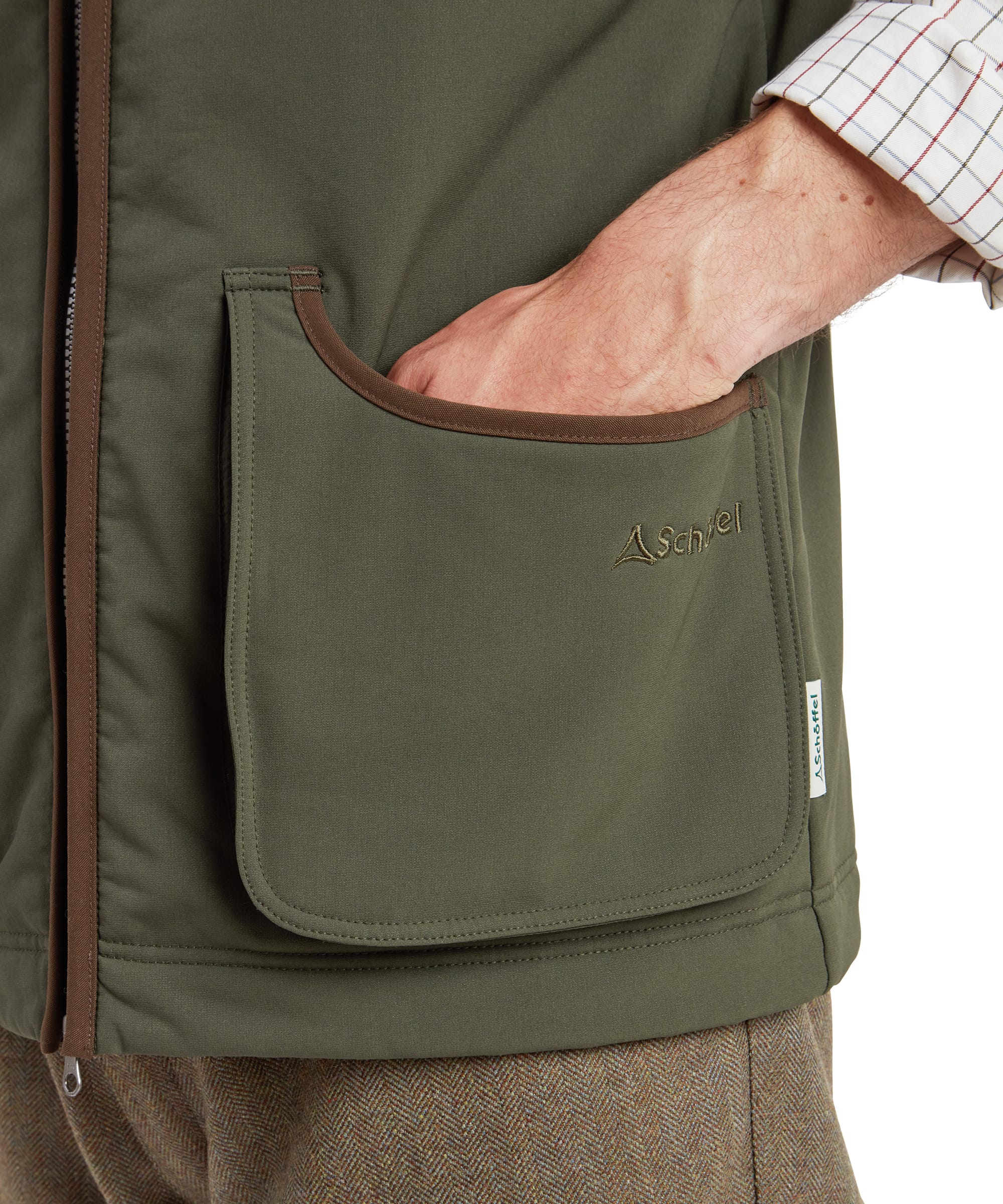 Schöffel Men's Gunby Gilet in Green — close-up of deep lower pocket showing Schöffel embroidered logo and suede trim edging, with check shirt sleeve visible