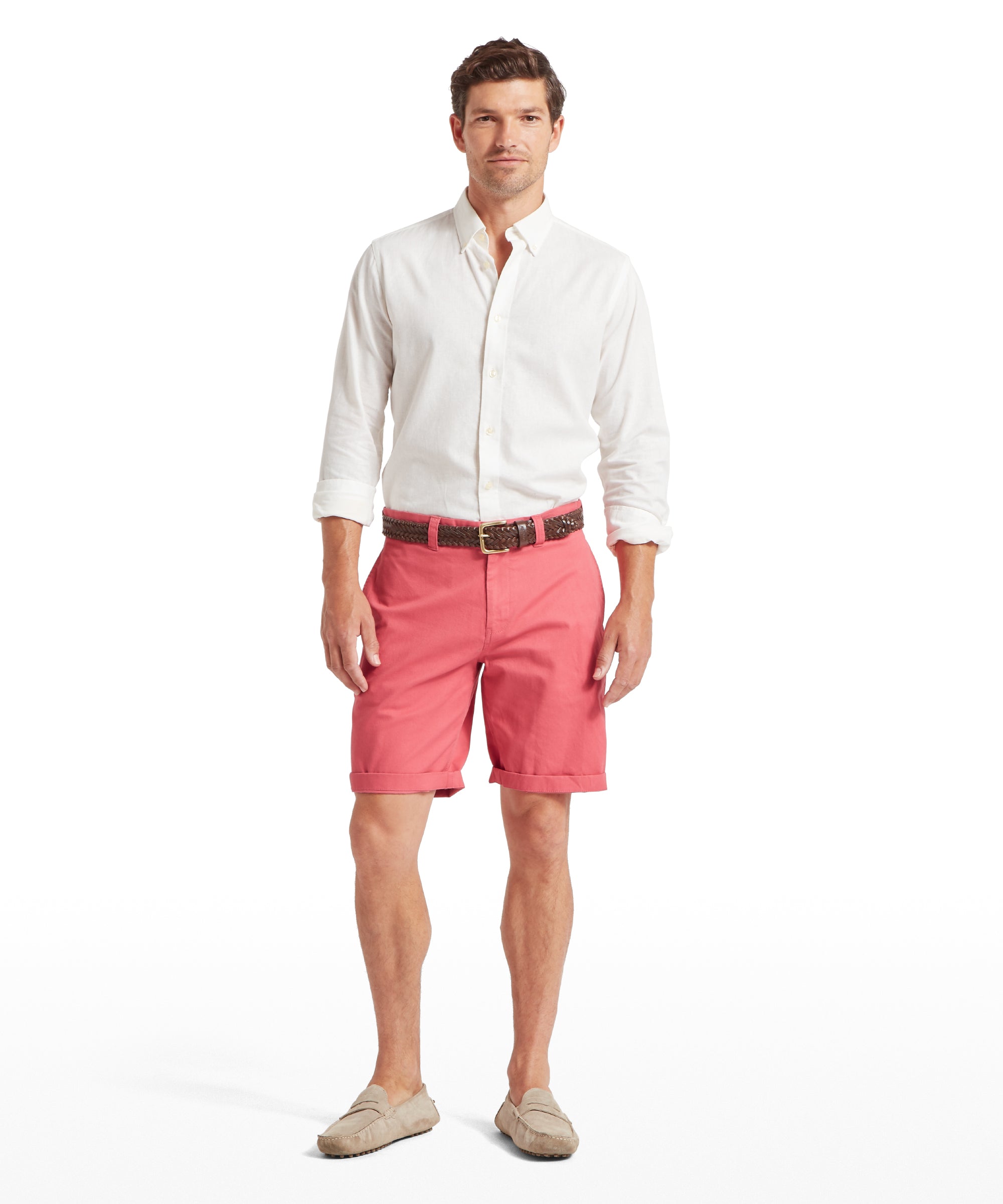 Man wearing coral pink shorts with a woven belt featuring a brown buckle. Paired with a white long-sleeved button-down shirt with rolled sleeves and light beige loafers. Standing front-facing against a plain white background.
