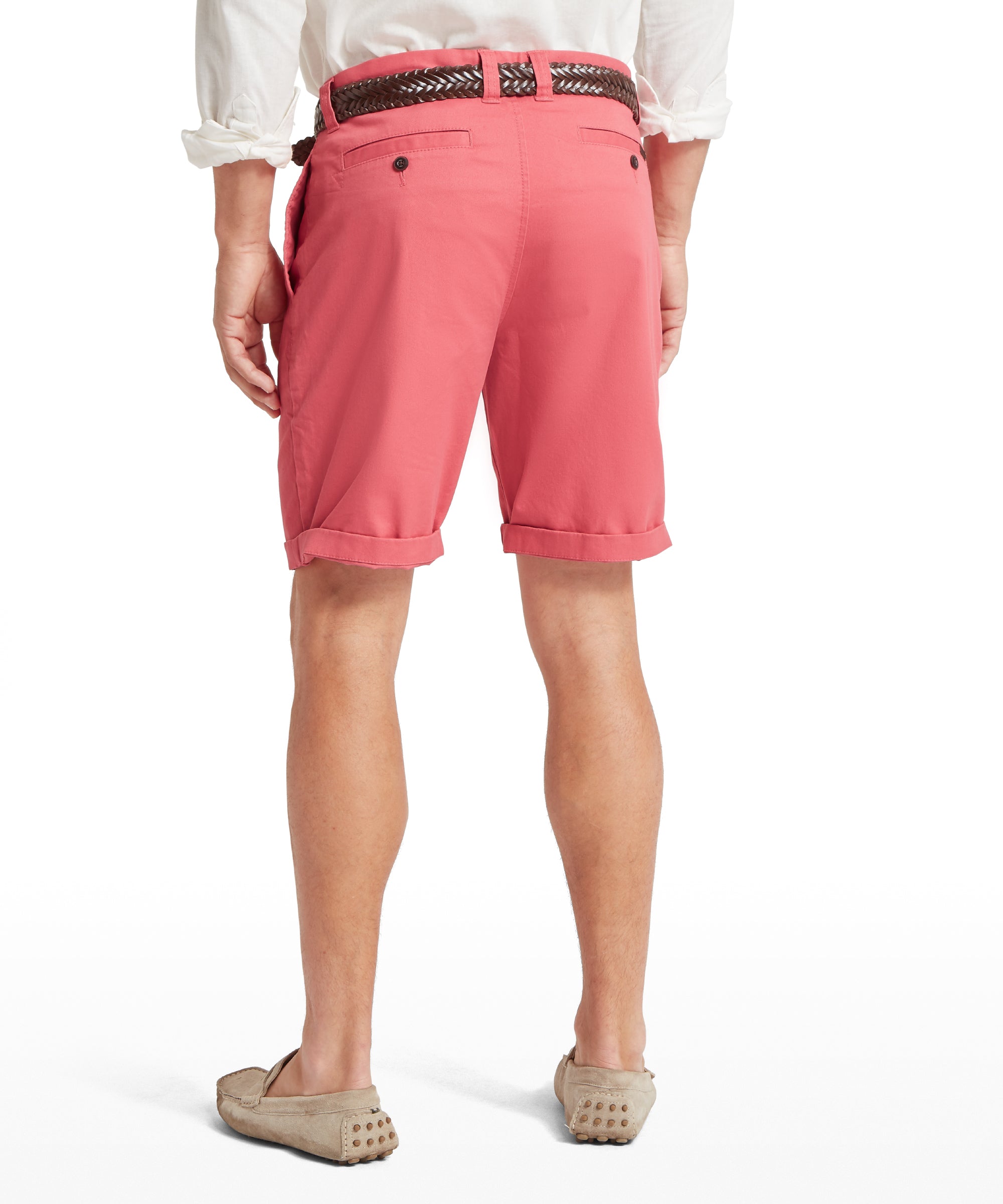 Back view of a man wearing coral pink shorts with a cuffed hem and two buttoned back pockets. Paired with a white long-sleeved shirt with sleeves rolled up, a dark brown woven belt, and light brown loafers with rubber stud soles. Standing upright against a plain white background.